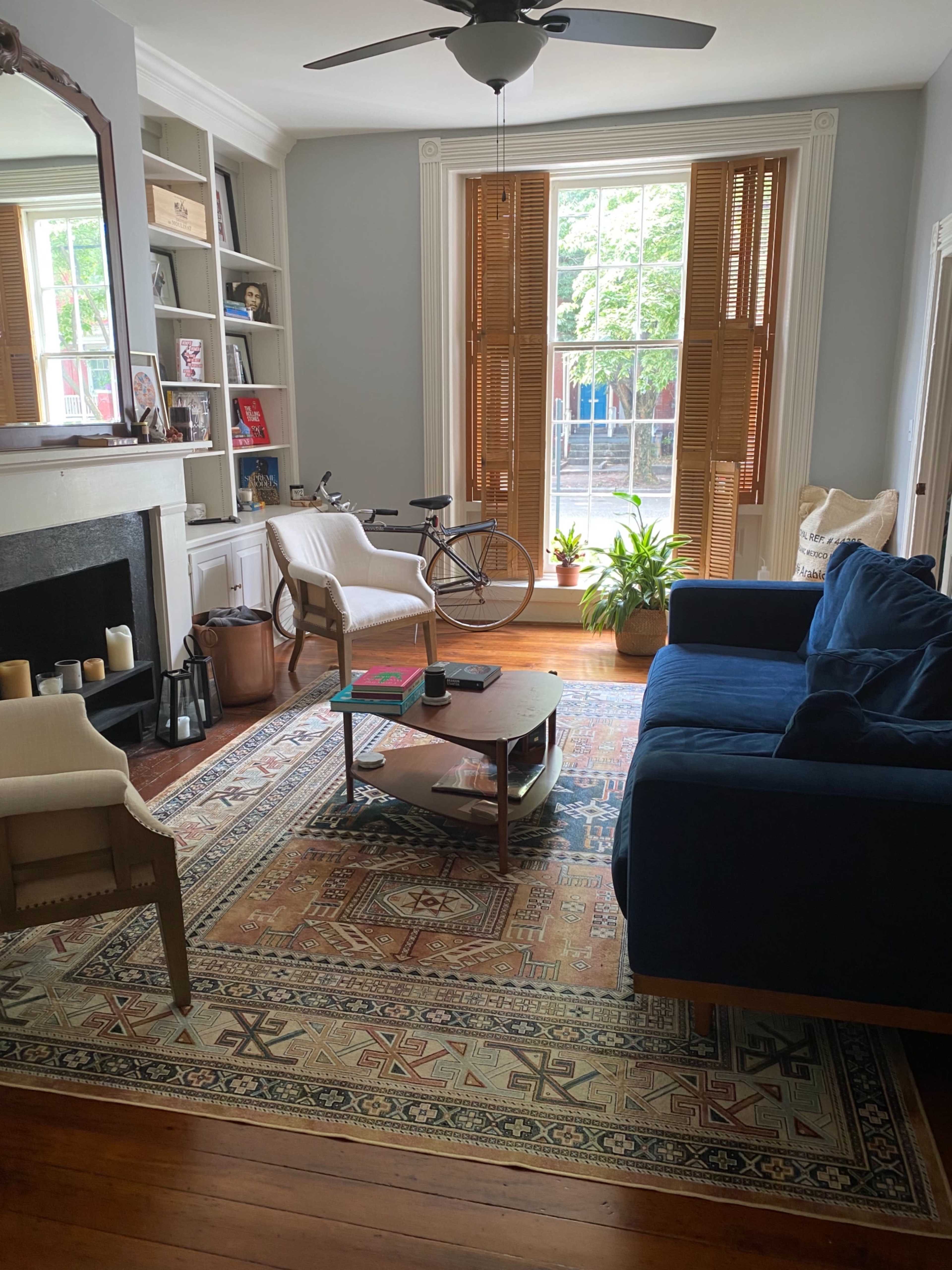 The living room features a blue sofa, a coffee table, a bicycle, and large windows with wooden shutters, all arranged around a patterned area rug.
