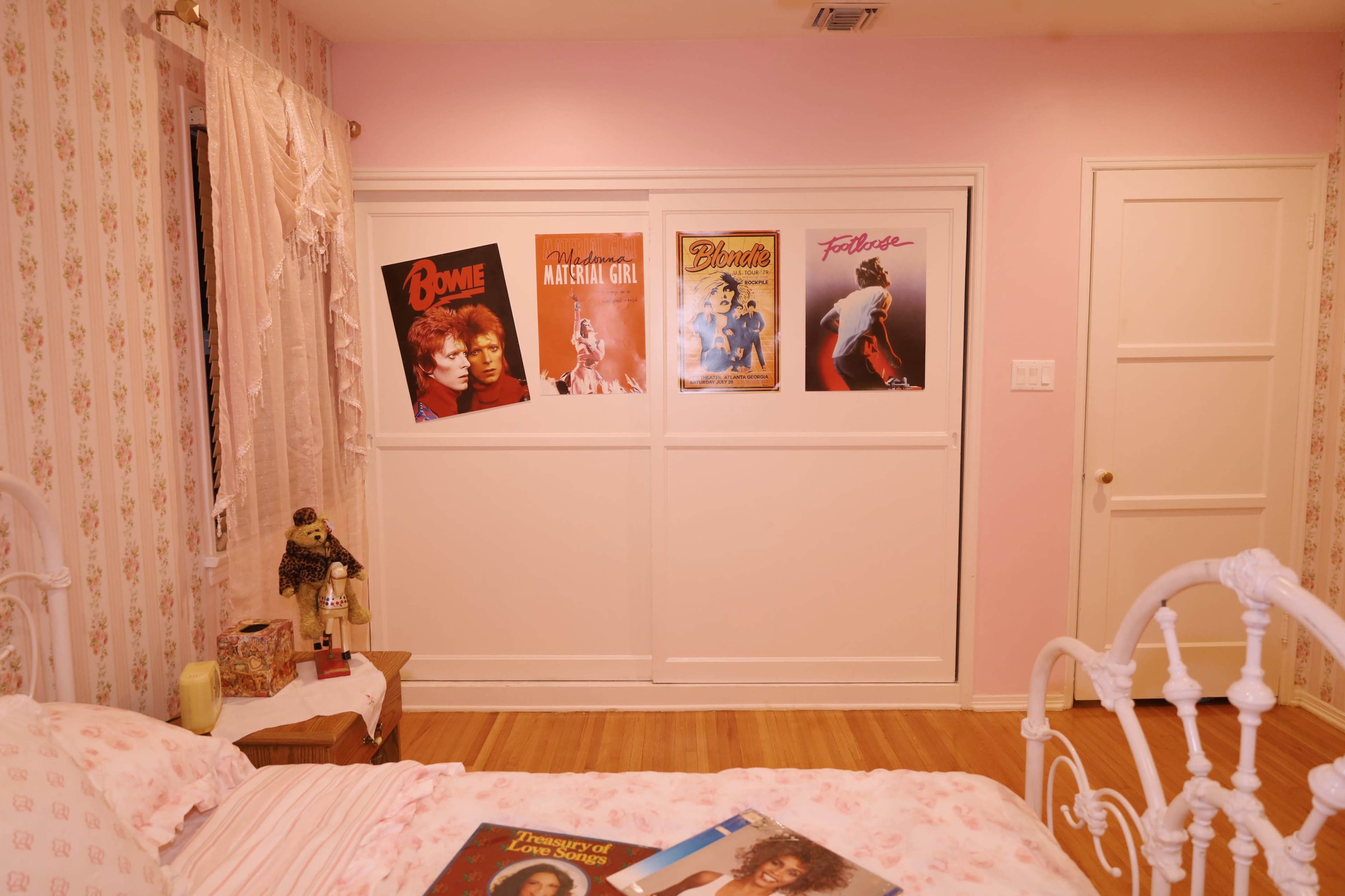 The image shows a pastel-colored bedroom with floral wallpaper, featuring a bed with a patterned blanket and posters mounted on the wall.