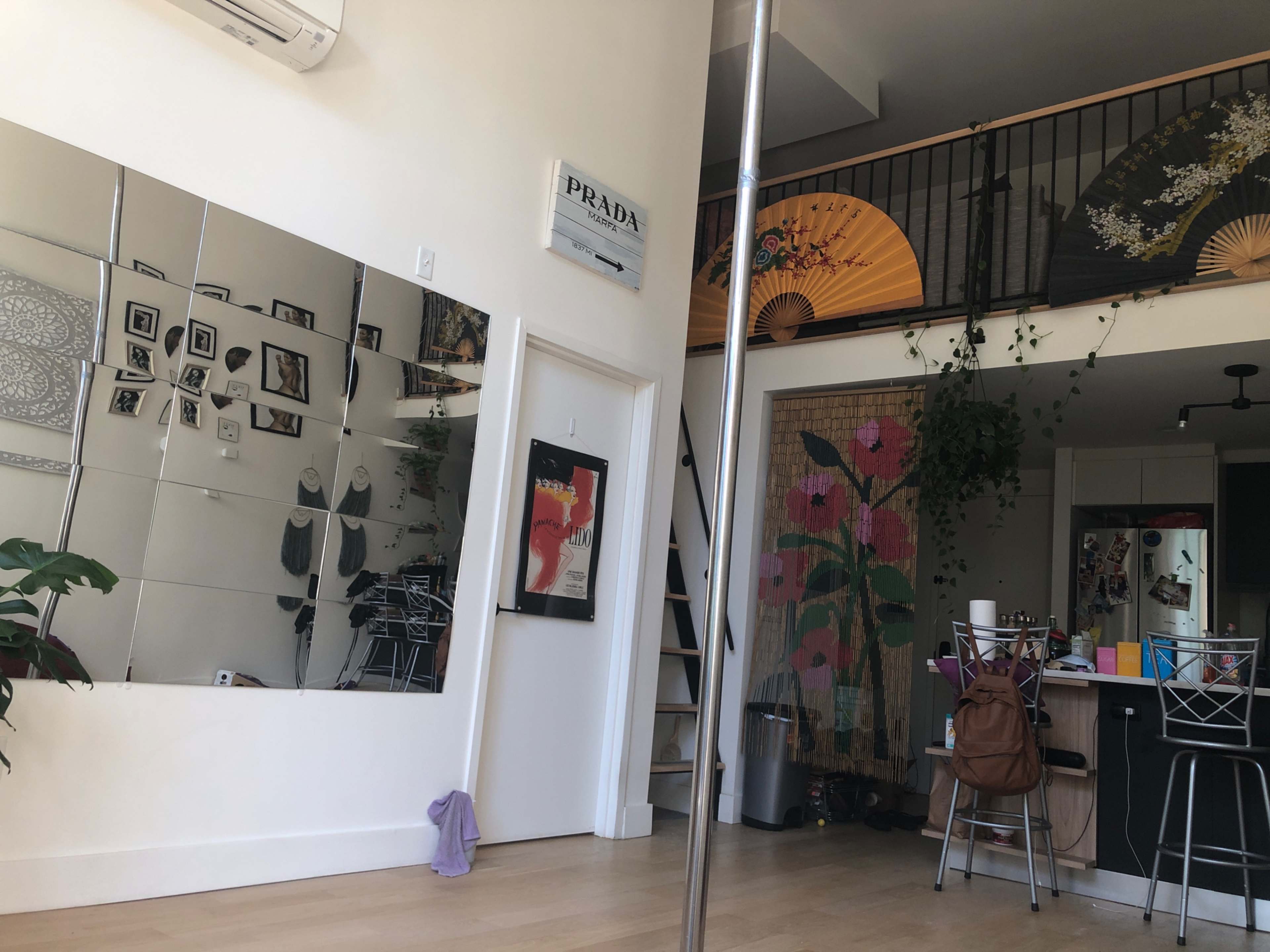 A spacious room with a pole in the center, a mirrored wall on one side, and artwork, plants, and furniture in the background.