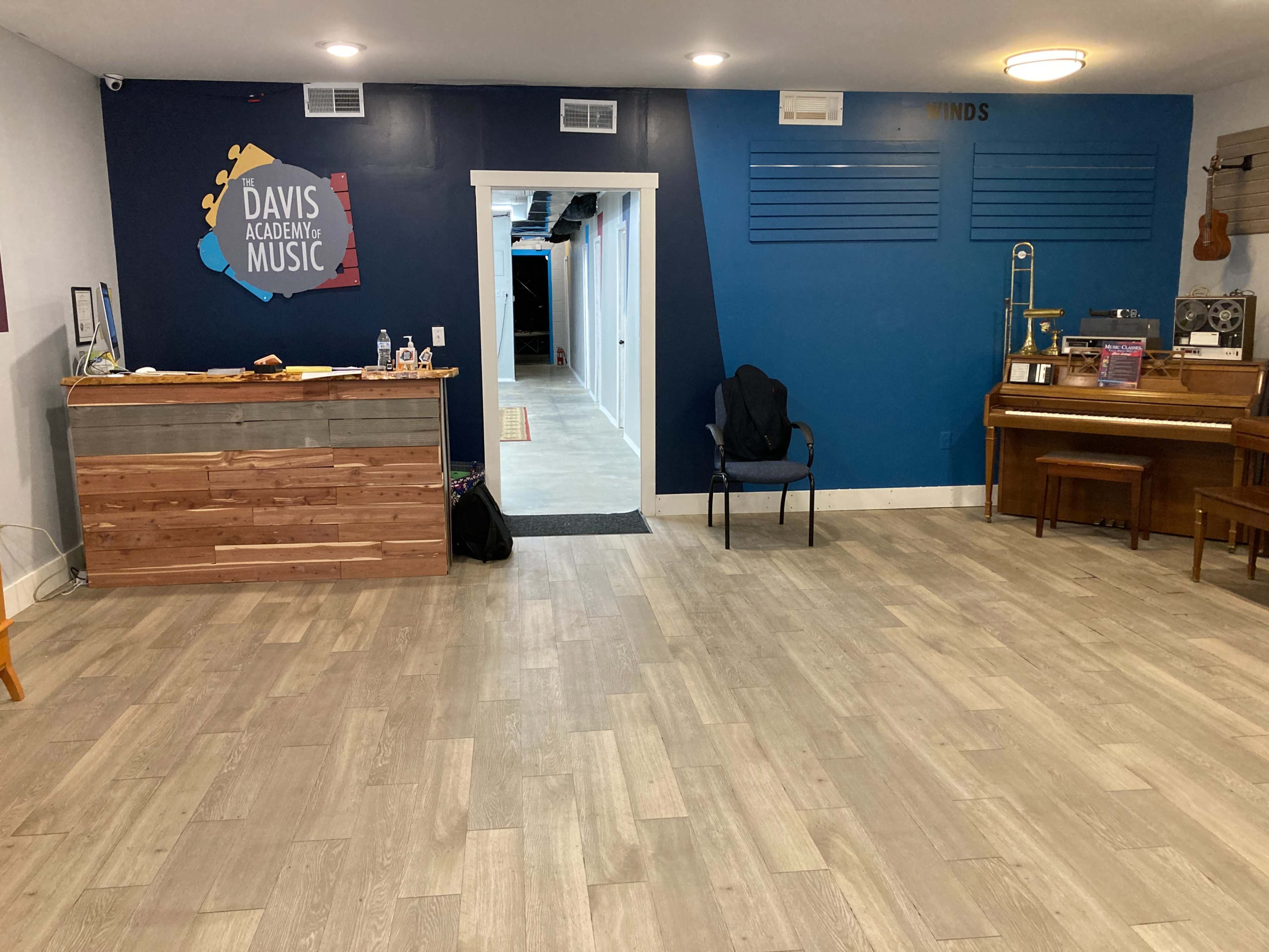 The image shows a music academy reception area with a wooden counter, a chair, a piano, and a blue accent wall featuring the academy's logo.