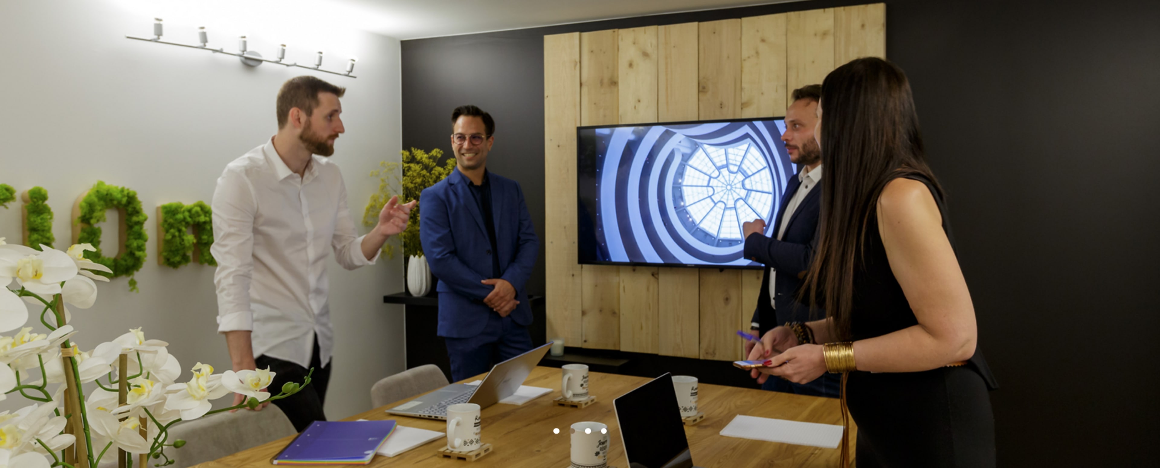 A group of four professionals is engaged in a discussion around a conference table, with a large screen displaying a circular design in the background.