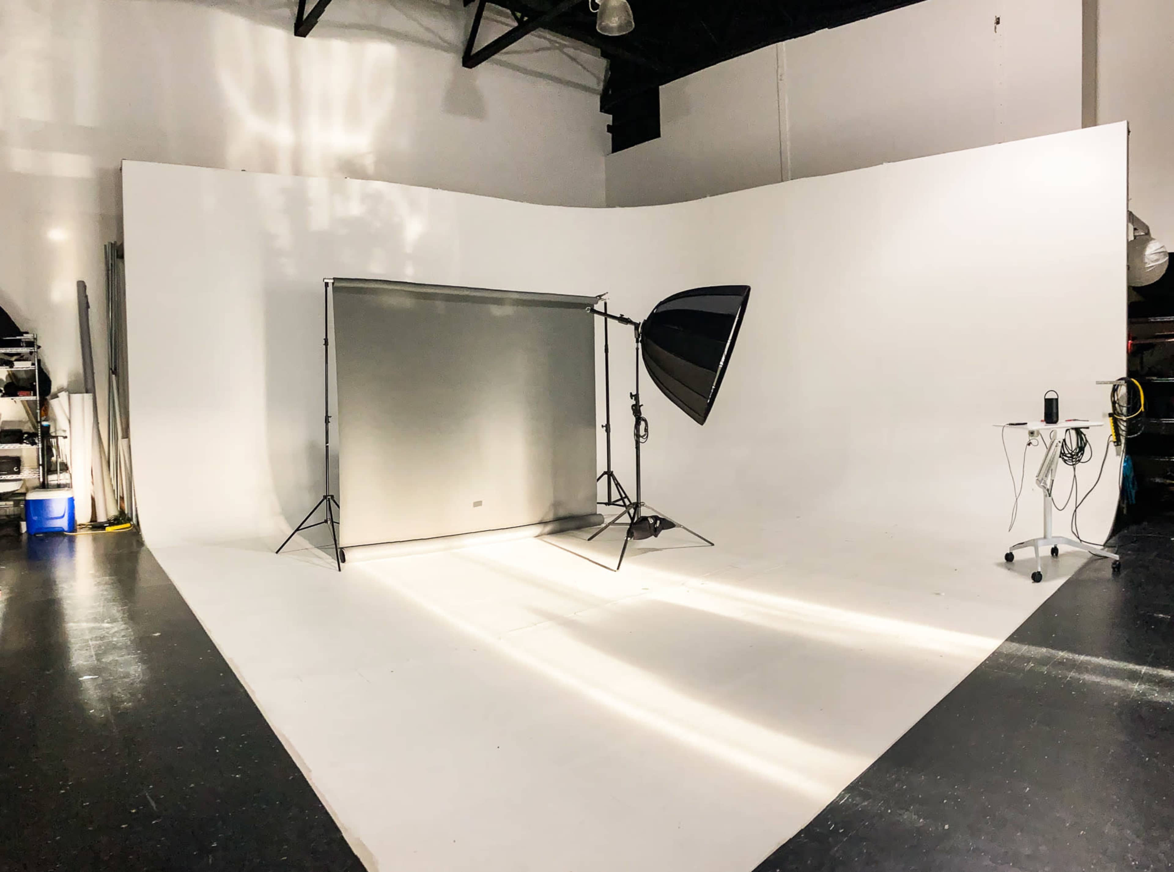 A photography studio with a large, white backdrop, a light source on a stand, and a small table with equipment.