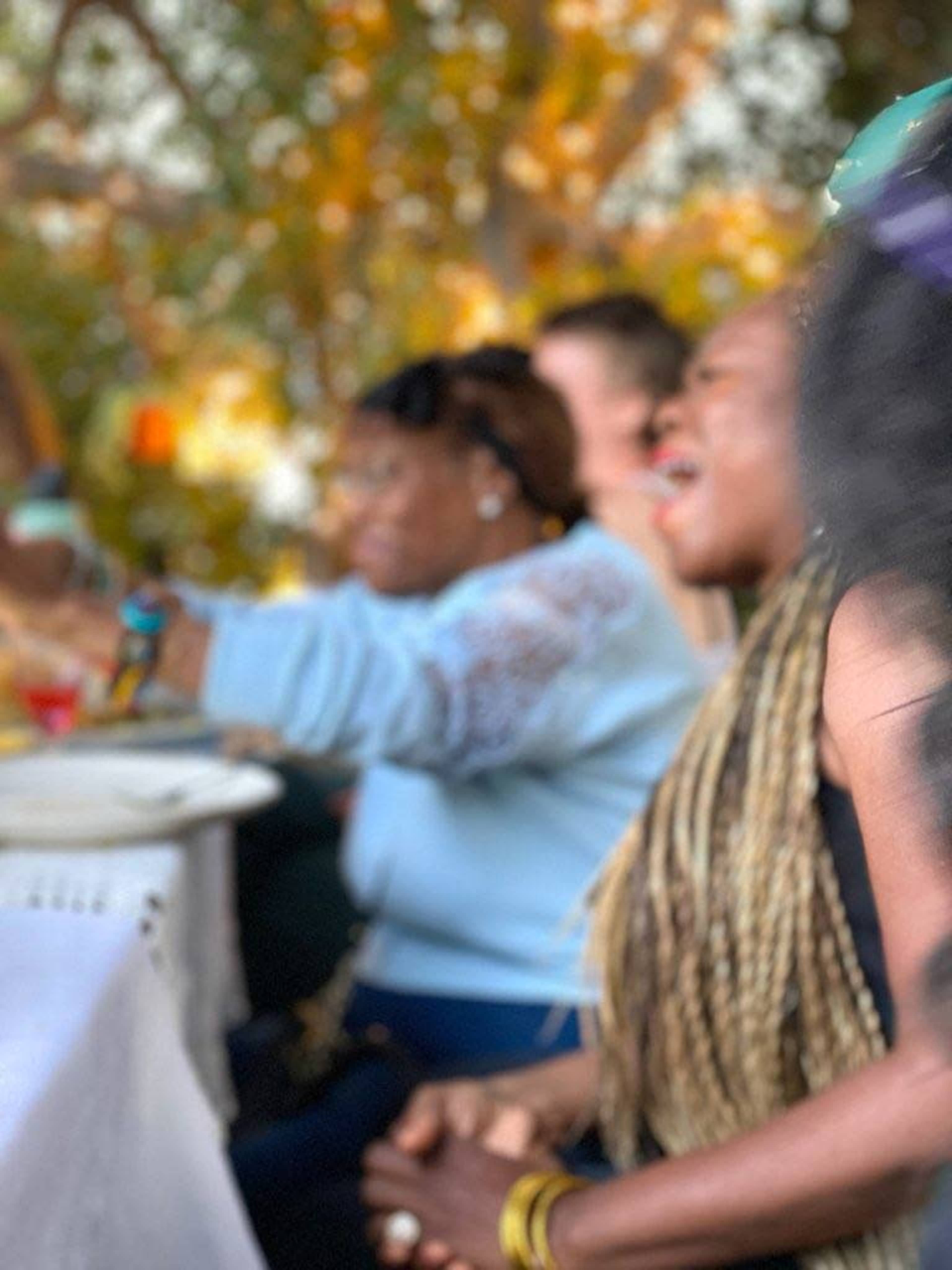 A group of people is gathered around a table, with one woman singing and others engaged in conversation, set against a backdrop of trees.
