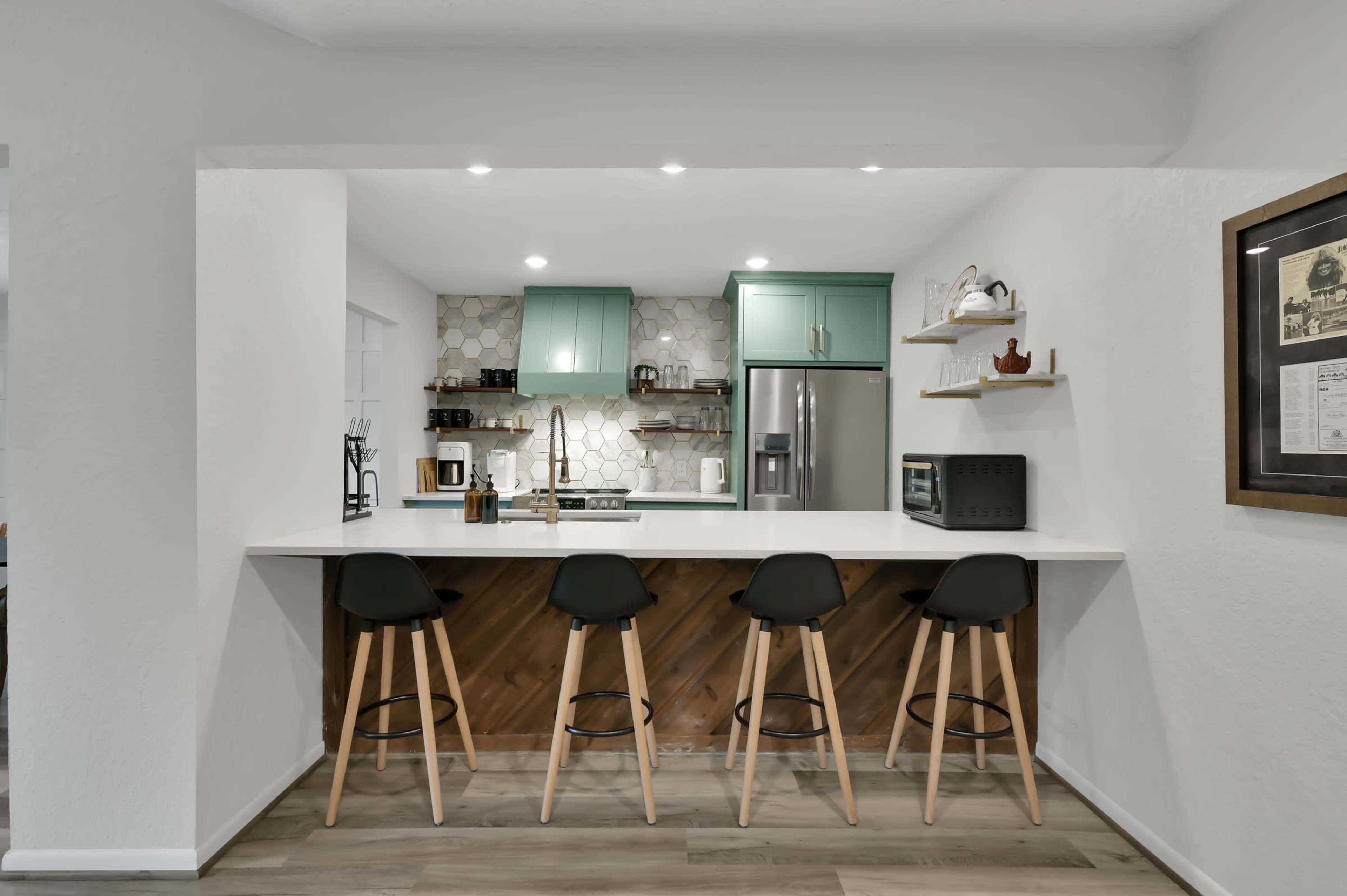 A modern kitchen features light green cabinetry, a white countertop, and wooden accents, with three stools arranged at a bar counter.