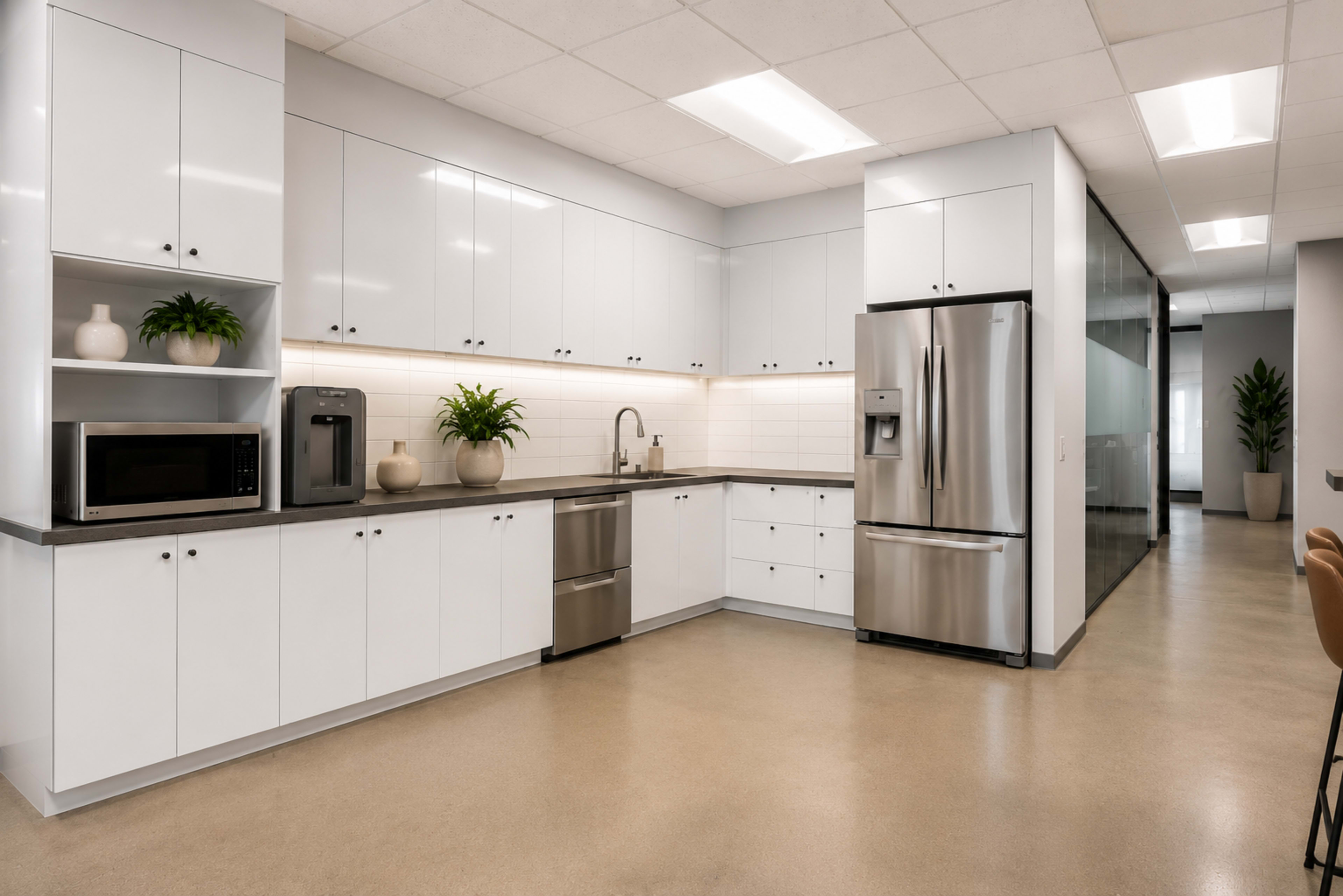 The image shows a modern kitchen area with white cabinetry, stainless steel appliances, and countertop plants in a well-lit office space.