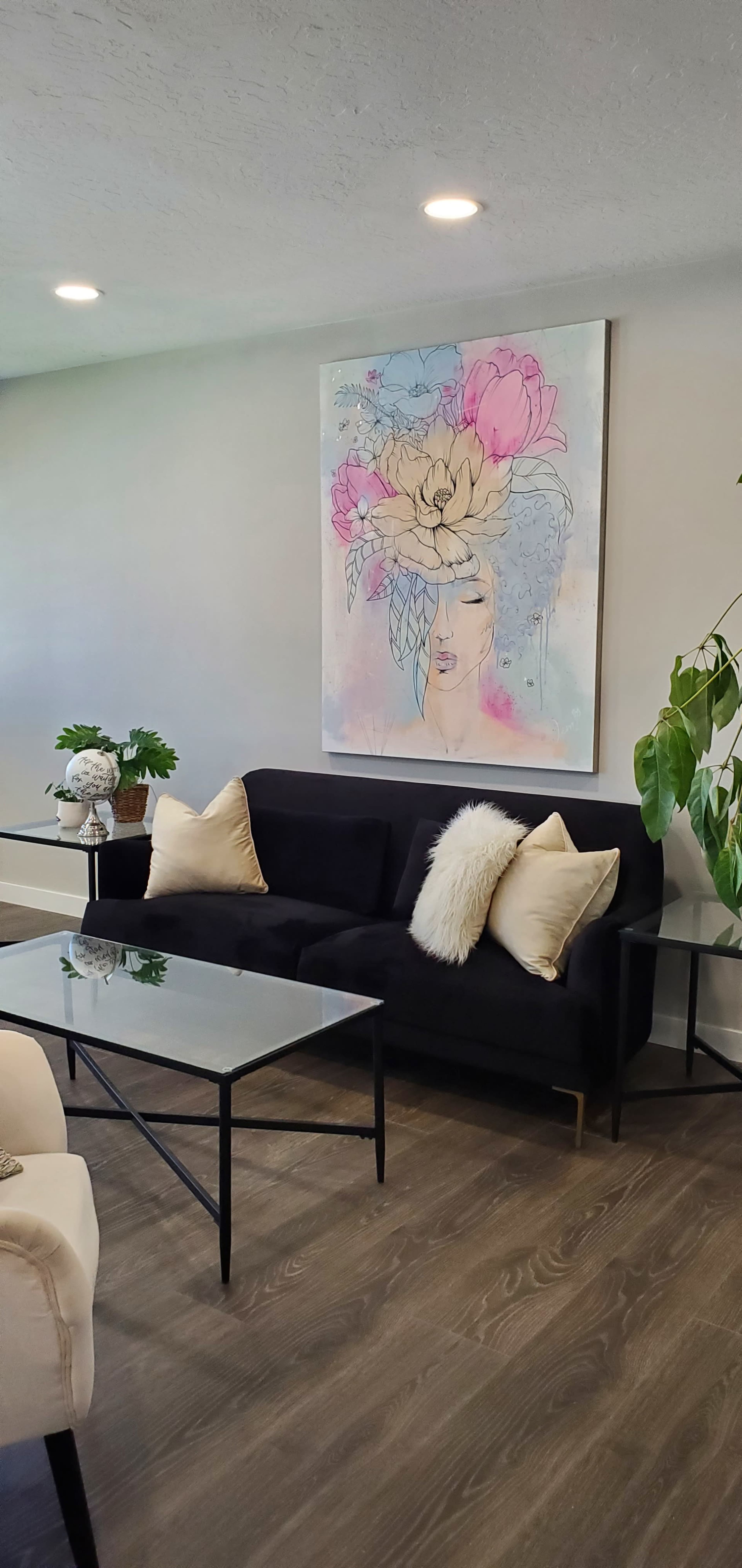 The living room features a black sofa with cream-colored pillows, a glass coffee table, and a large floral artwork hanging on the wall.