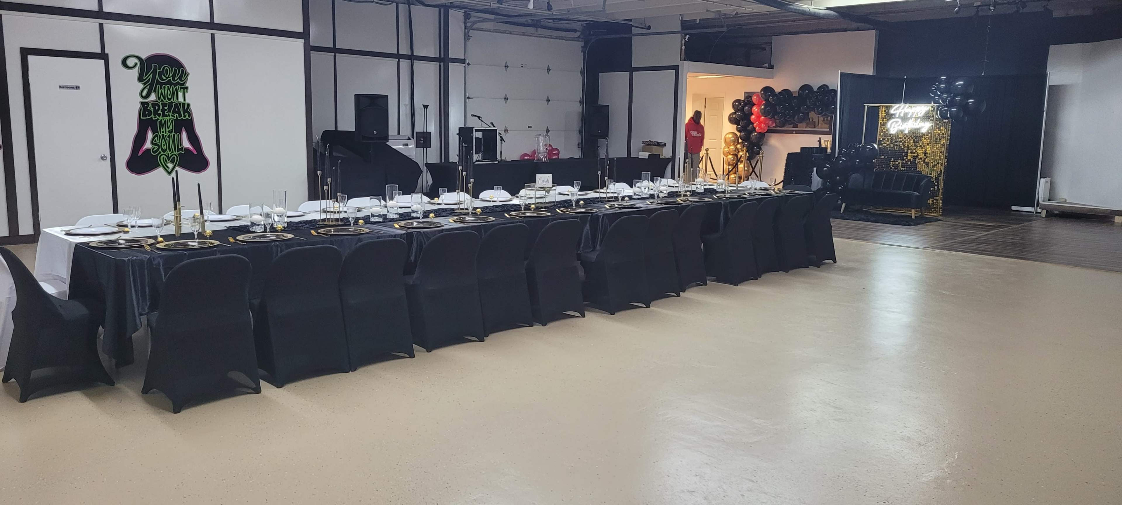 A long dining table set for a formal event is positioned in a spacious venue with black tablecloths and decorations.