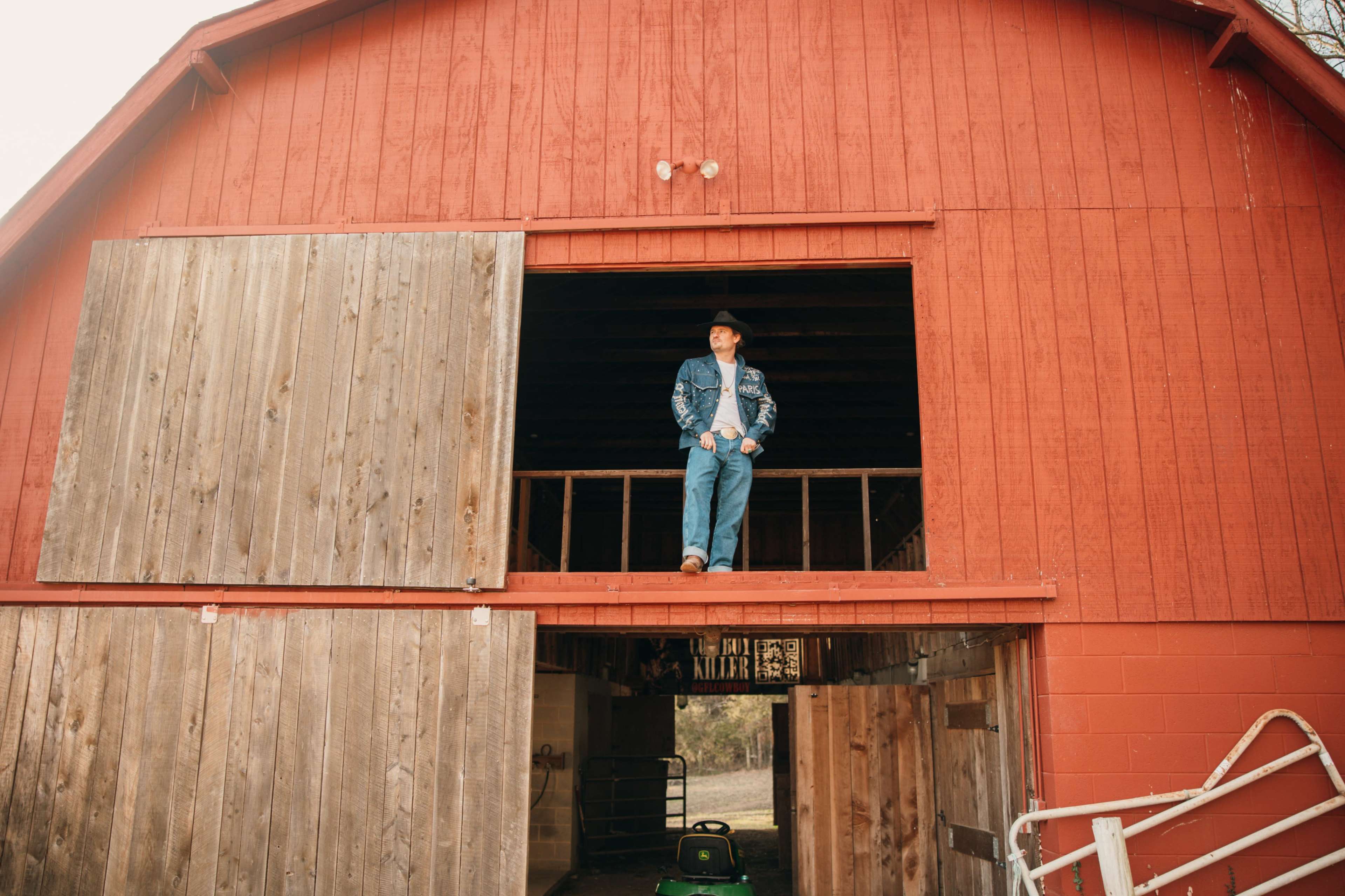 A man in a cowboy hat and denim clothing stands on the upper balcony of a red barn, overlooking the entrance below.