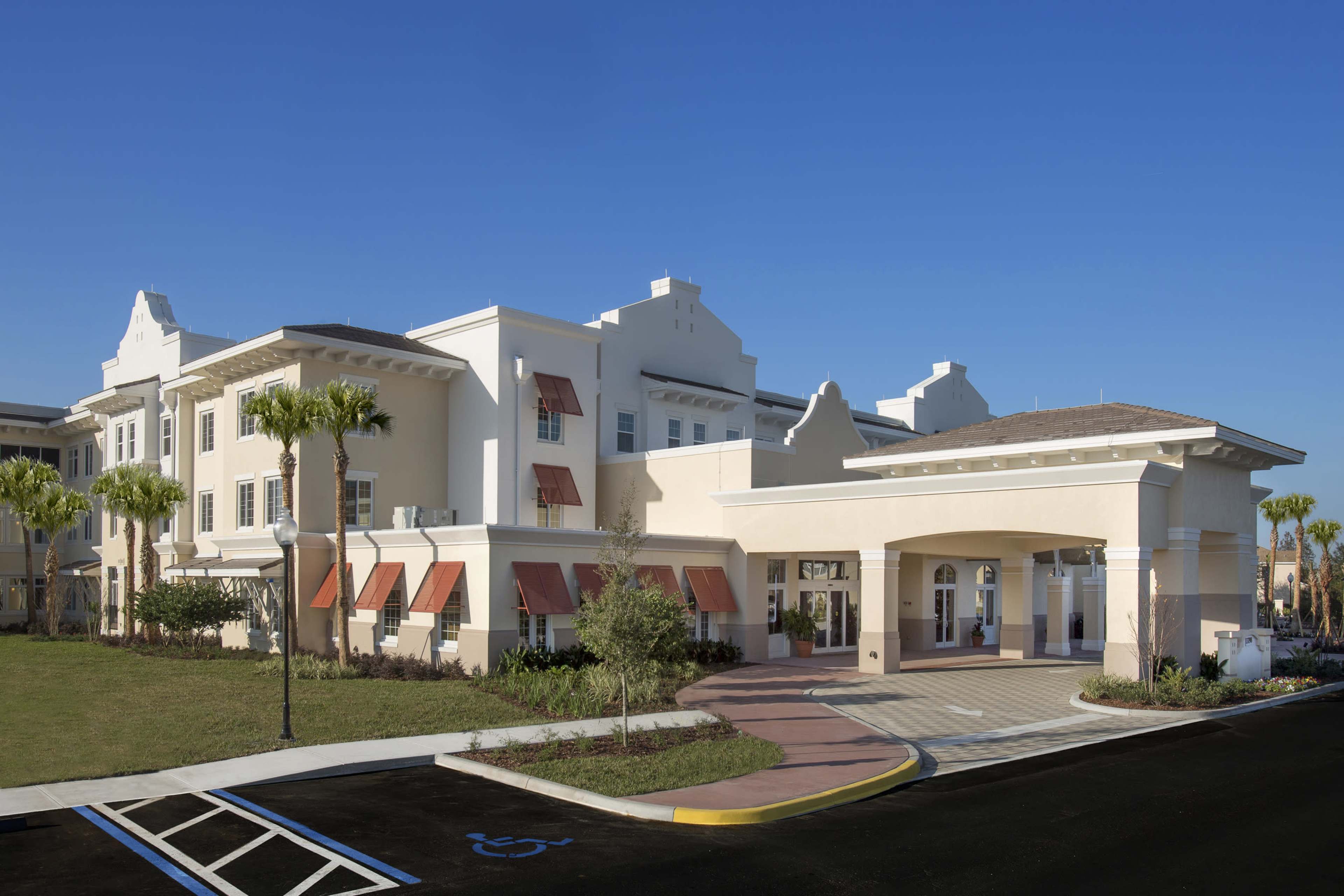 The image shows a large, two-story building with a light-colored exterior, featuring palm trees and a landscaped entrance with a circular drive.