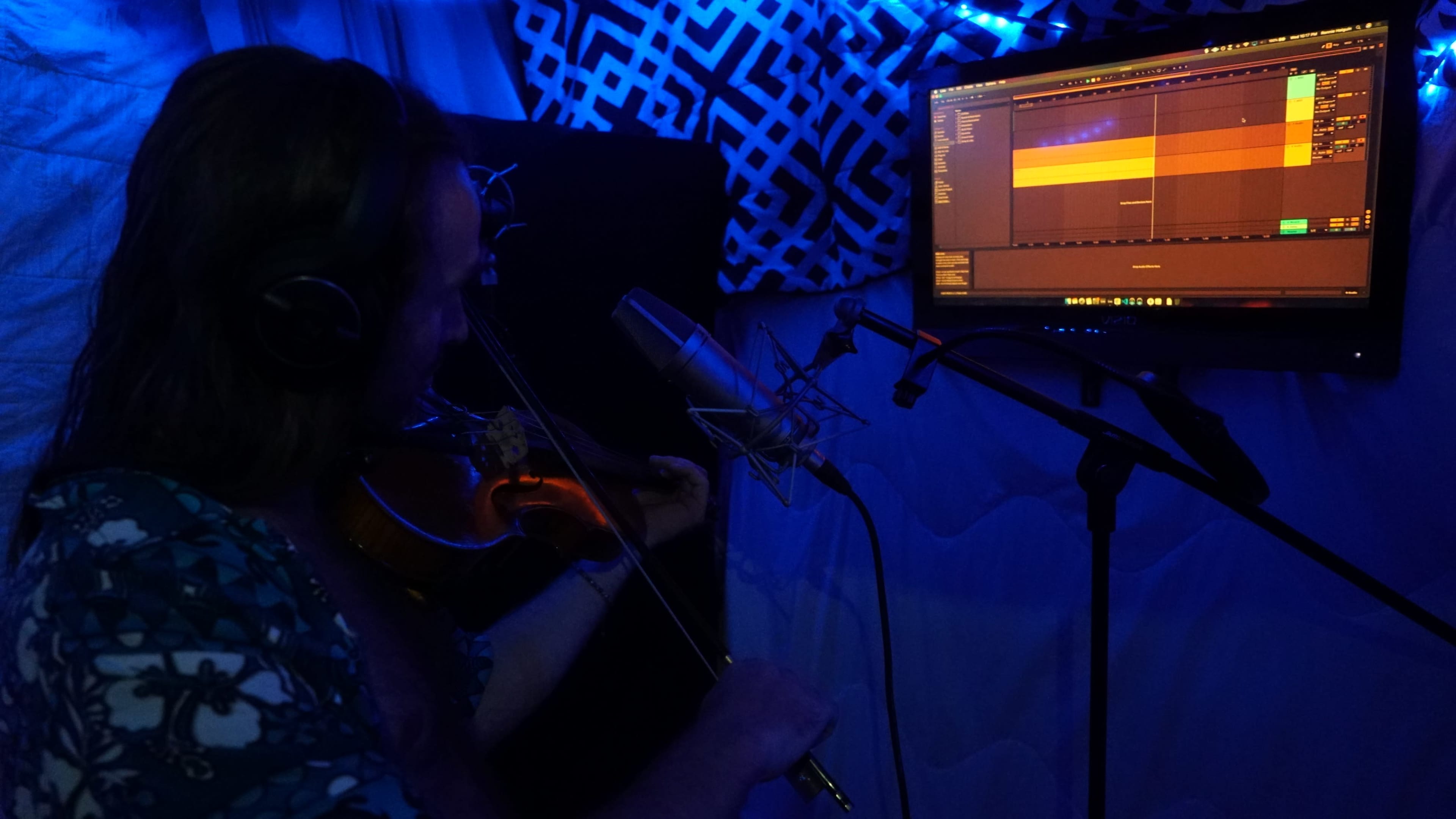 A musician plays the violin in a dimly lit recording studio with a computer screen displaying audio software.