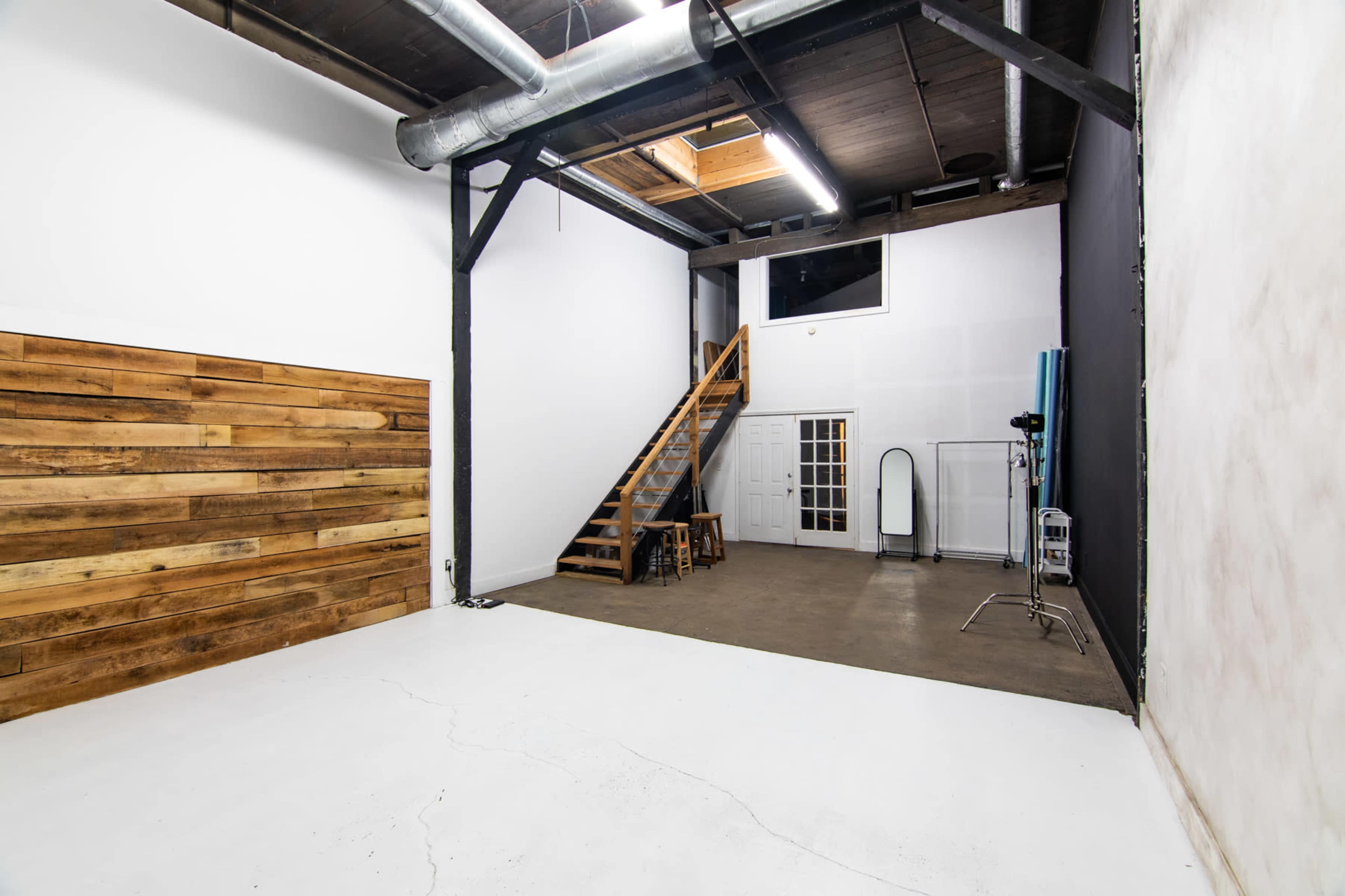 An empty studio space featuring wooden wall panels, a staircase leading to an upper level, and a large window for natural light.