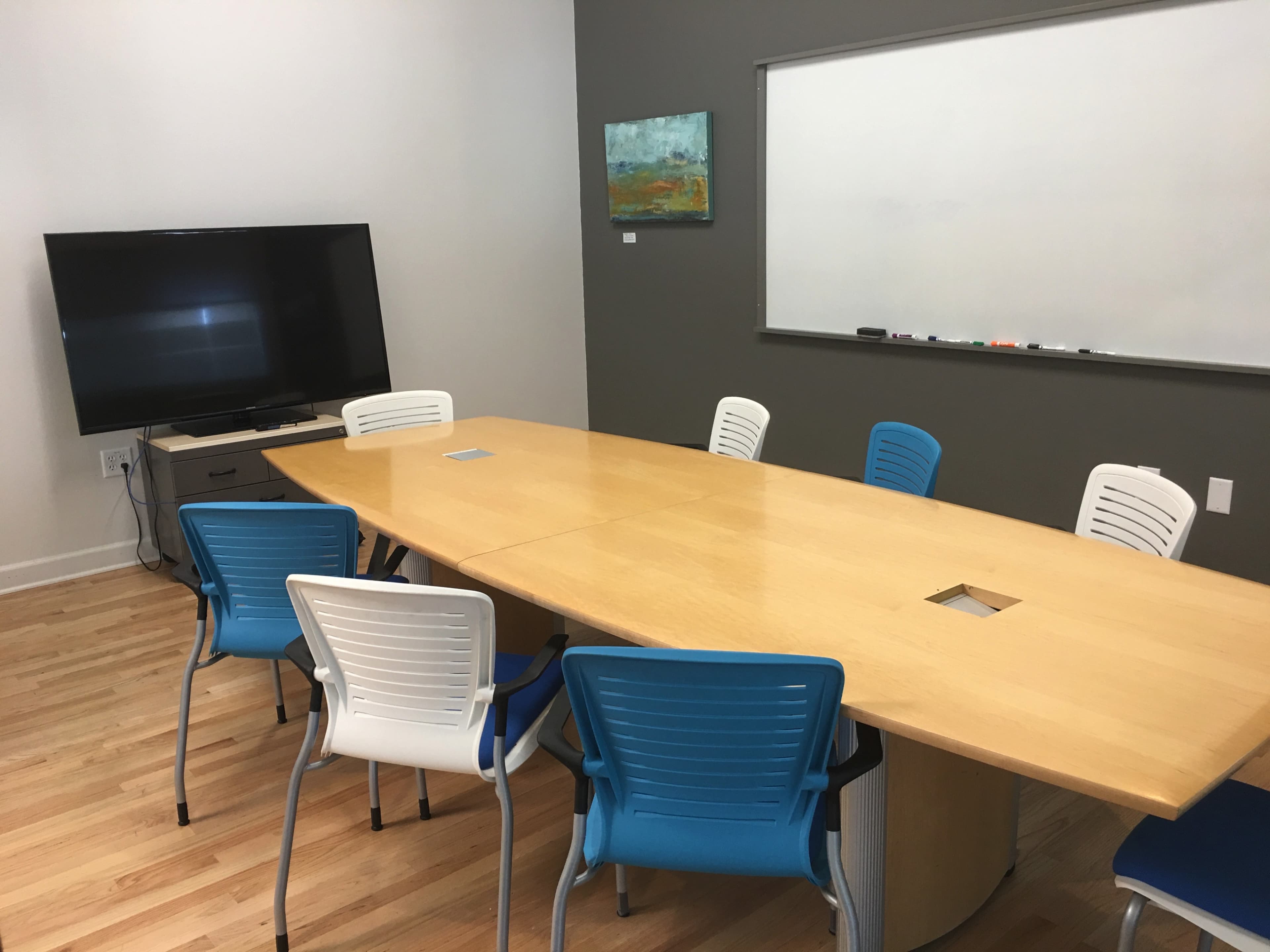 A wooden conference table surrounded by blue and white chairs, with a television and whiteboard on the wall.