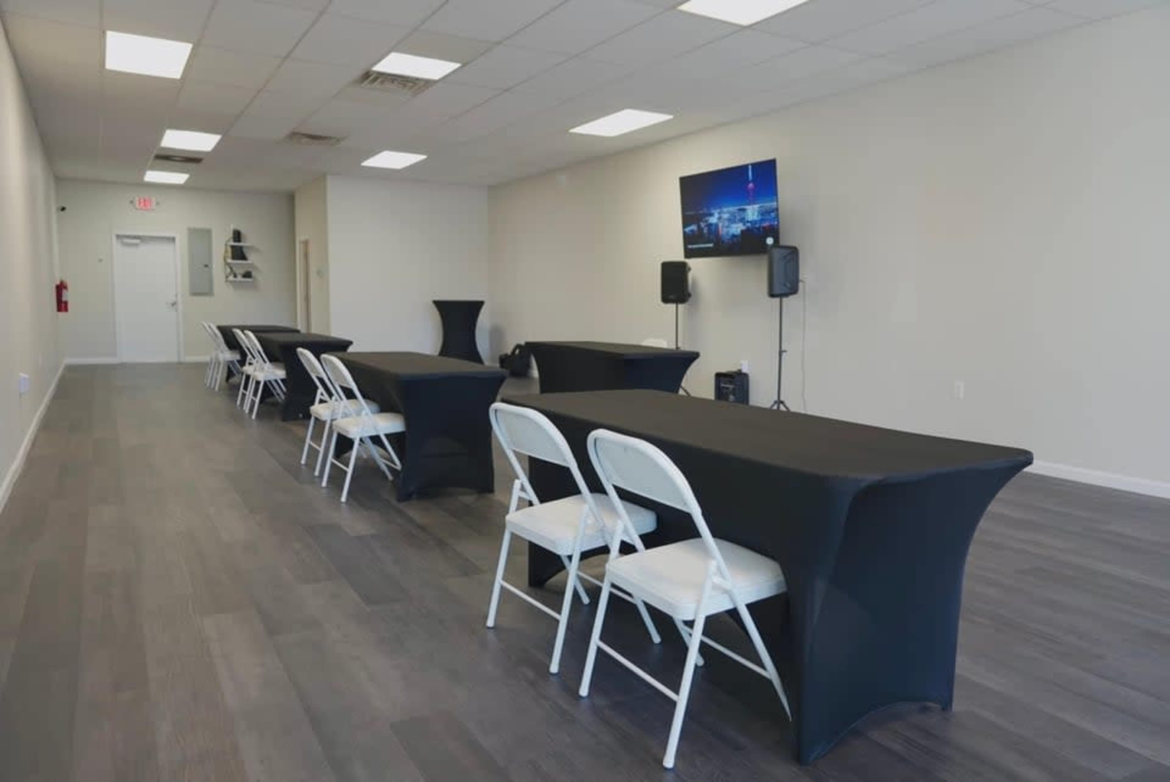 The image shows a spacious room arranged with several long tables covered in black tablecloths, accompanied by white chairs, and features a television and speakers at one end.