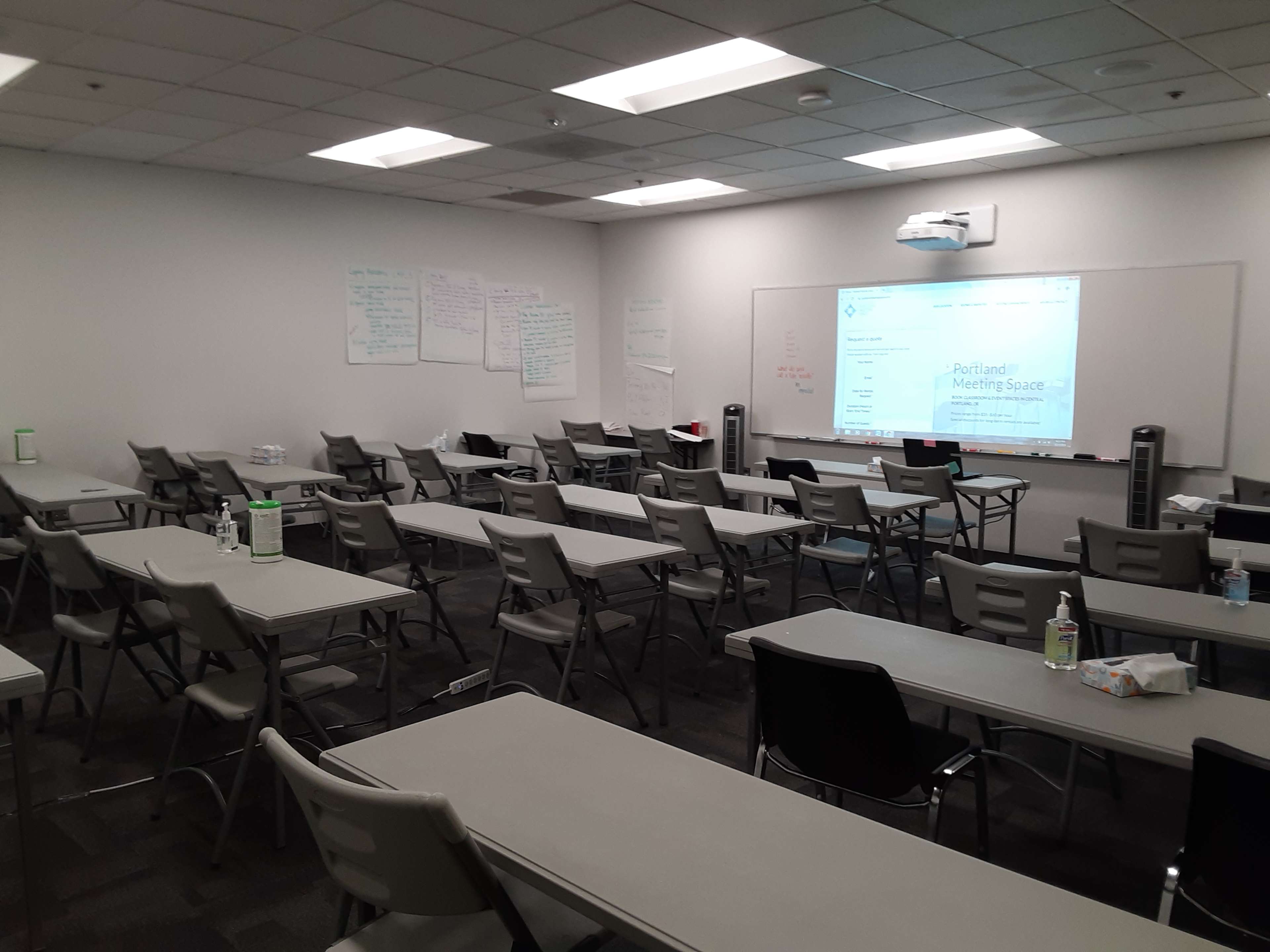 A meeting room is set up with rows of tables and chairs, featuring a projector screen displaying information about a Portland meeting space.