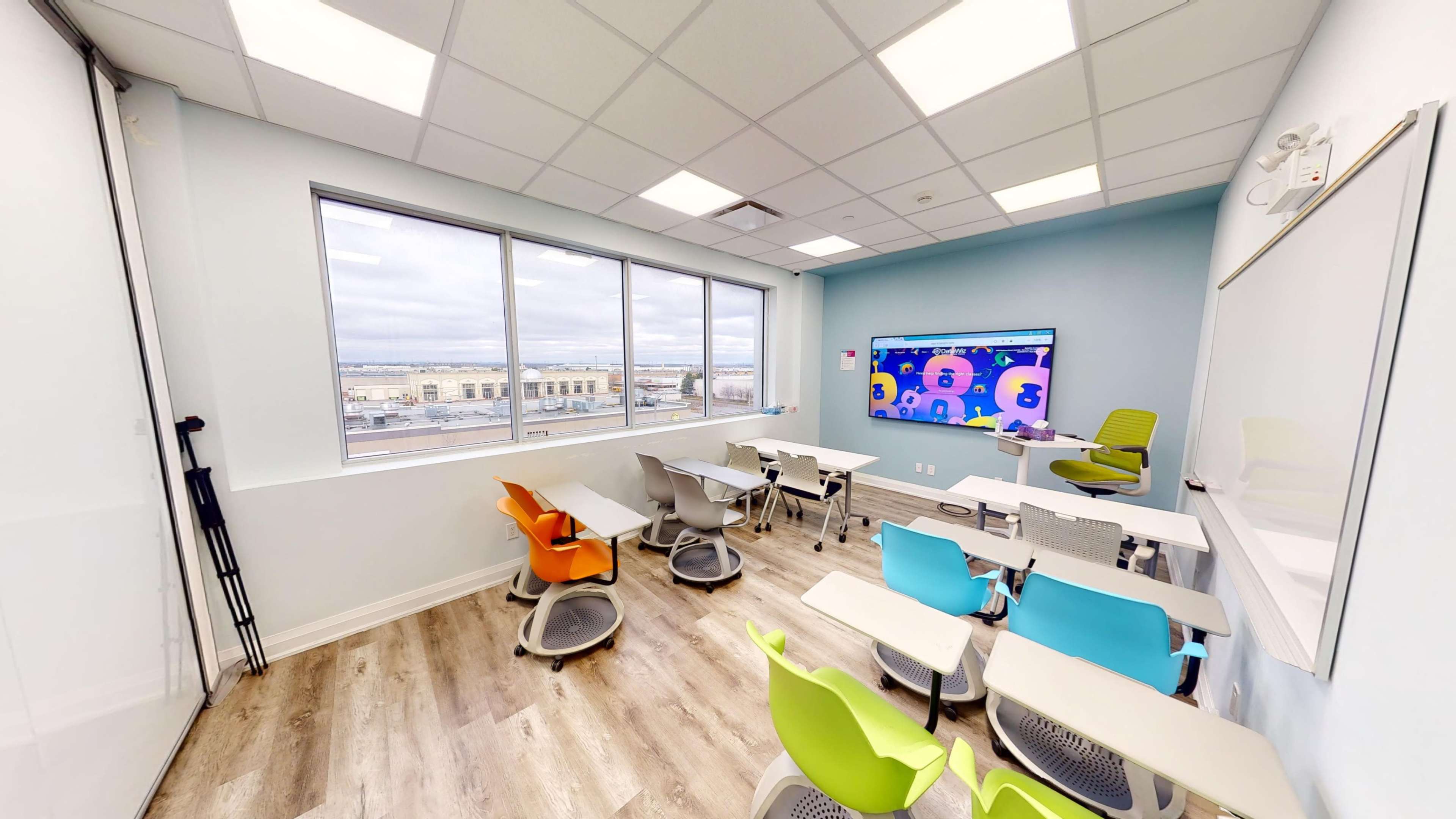 A modern classroom with colorful chairs arranged around small tables, equipped with a large screen displaying animated graphics and large windows offering an outside view.