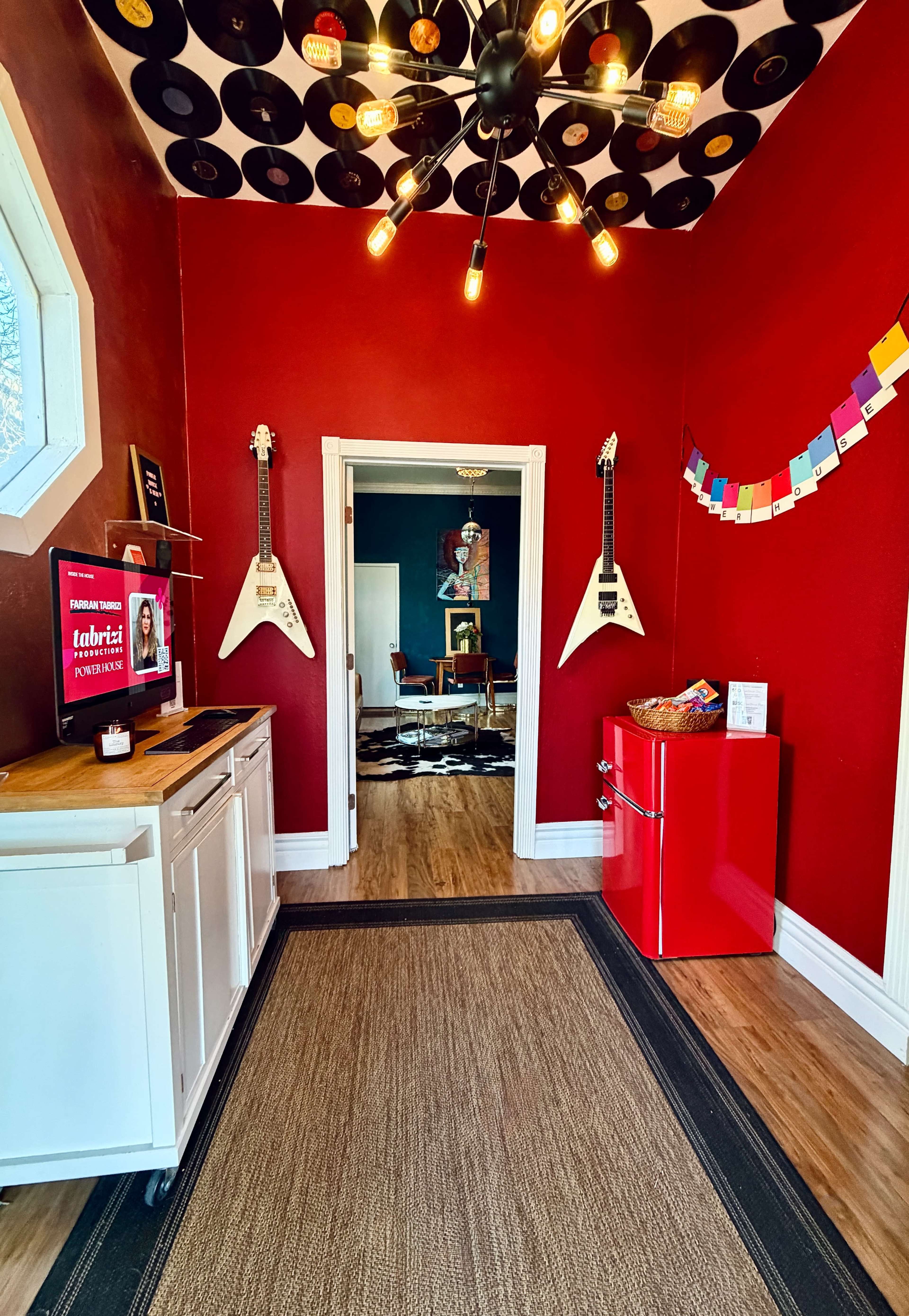 The image shows a vibrant red room featuring a vintage-style red refrigerator, two white electric guitars mounted on the wall, and a ceiling decorated with vinyl records.