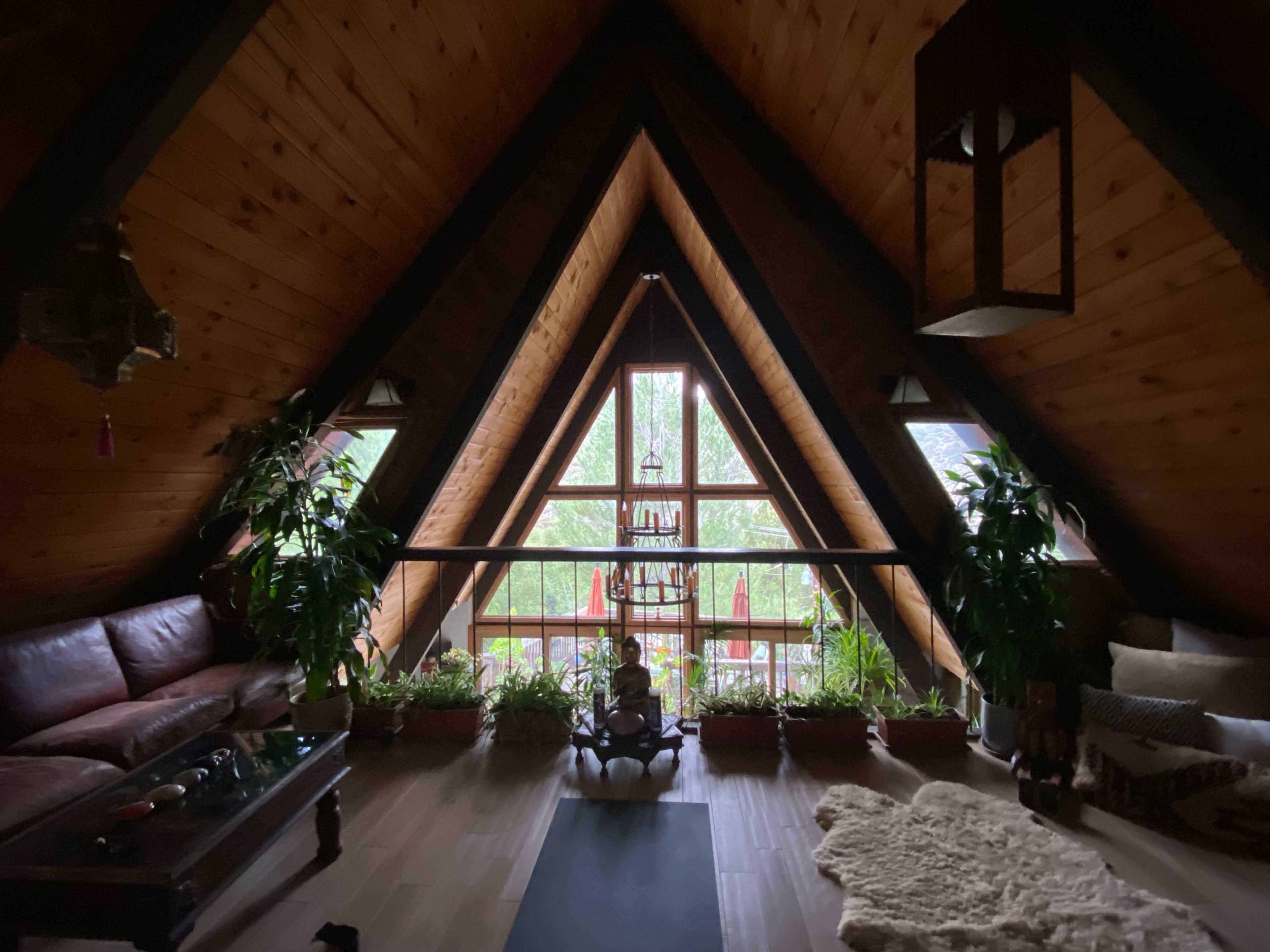 A cozy attic space with wooden walls, a triangular window view filled with greenery, and a seating area featuring a brown couch and indoor plants.