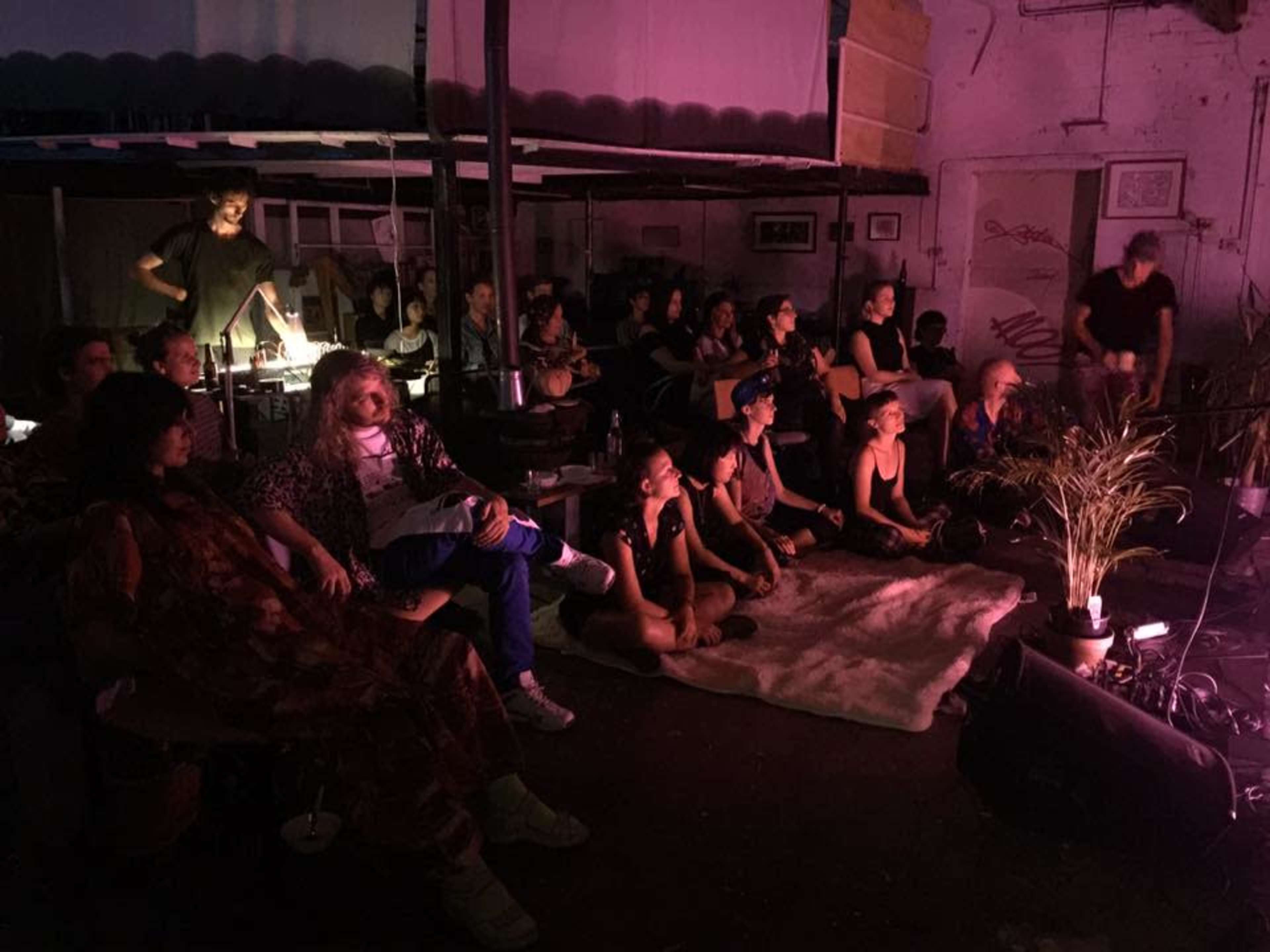 A group of people is seated in a dimly lit room, some facing a performance area while others appear engaged in conversation.