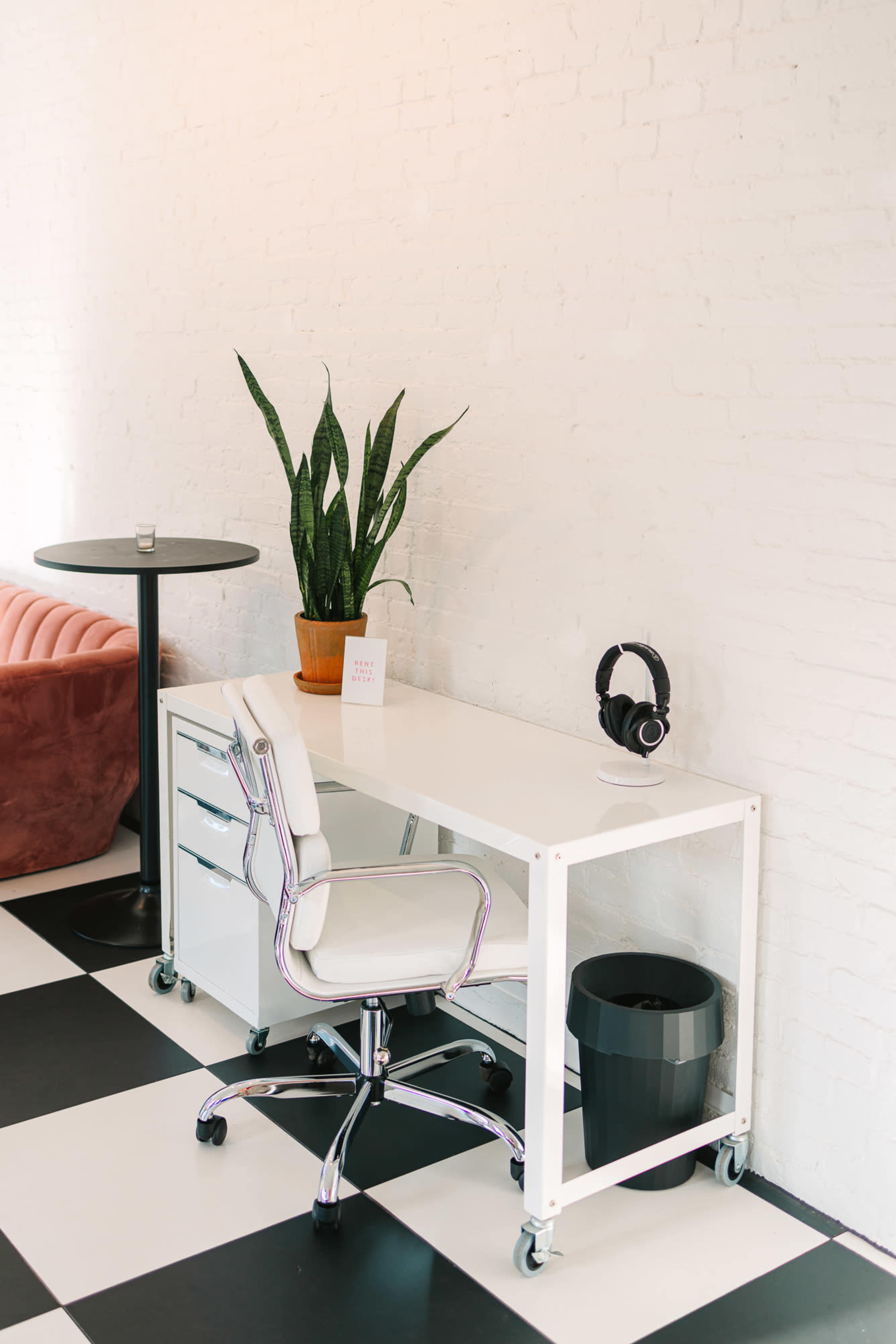 A modern workspace features a white desk with a rolling chair, headphones, a plant, and a small round table nearby, all set against a black and white checkered floor.