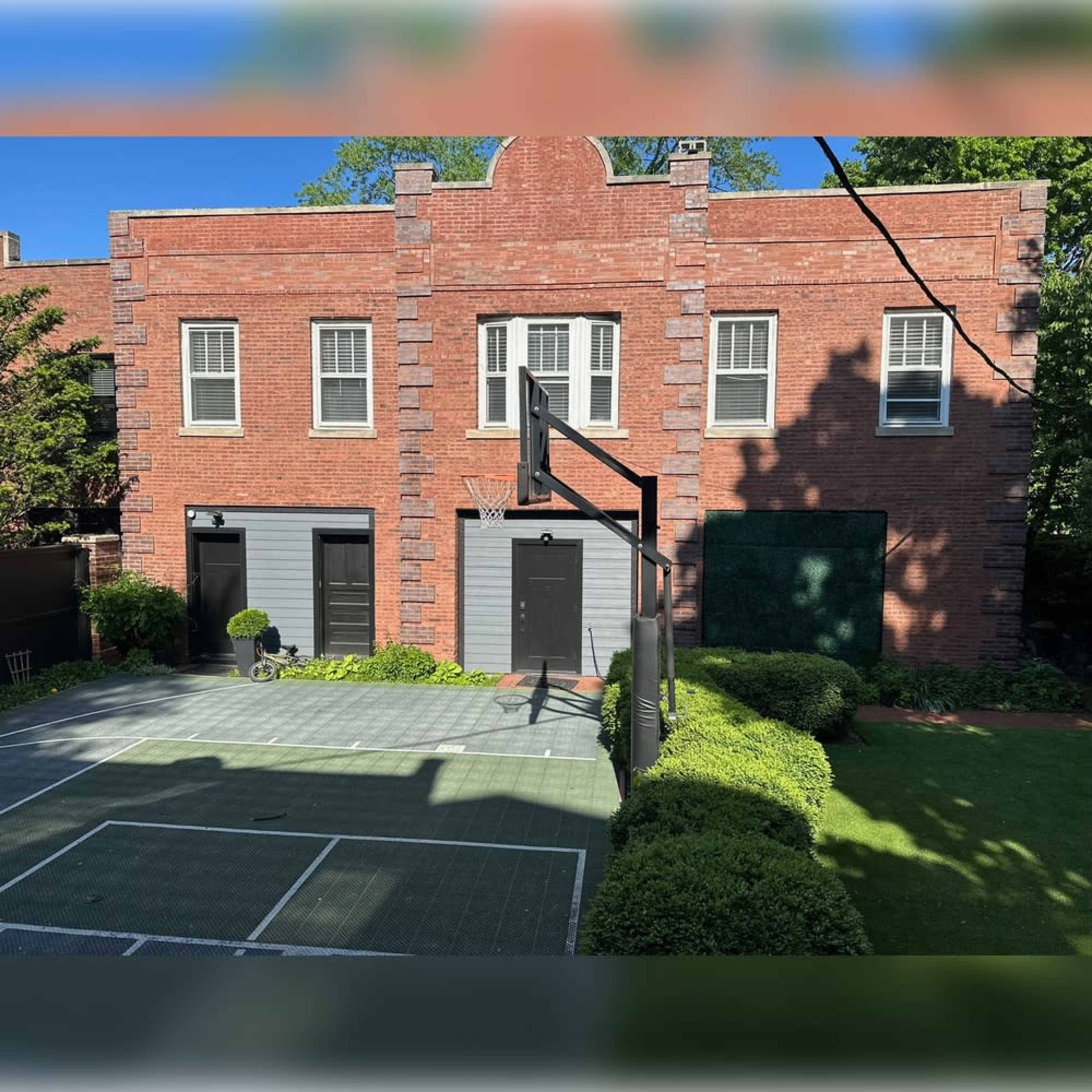 The image shows a brick building with multiple windows and doors, overlooking a basketball court surrounded by manicured shrubs and grass.