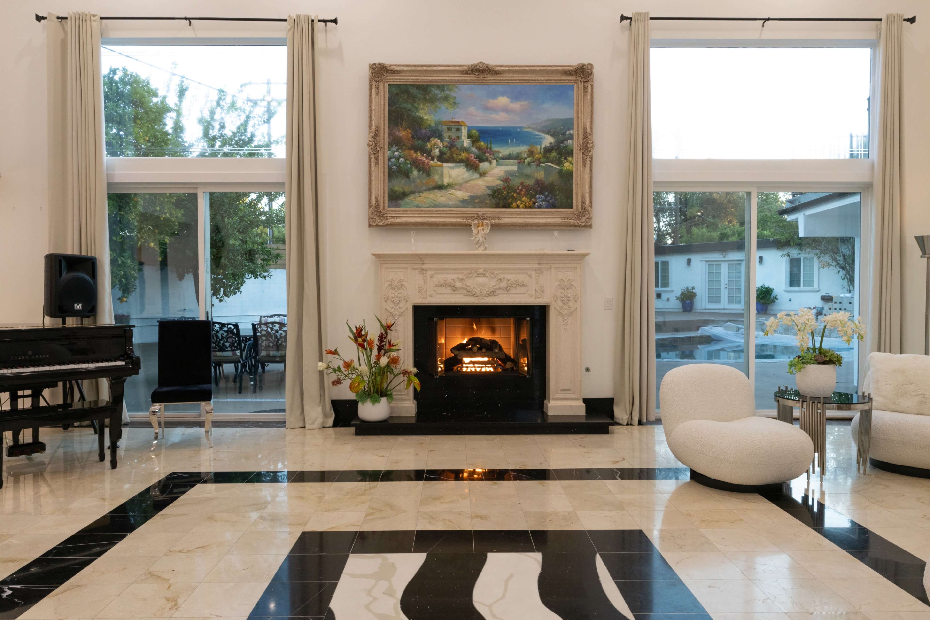 The image shows a spacious living room featuring a fireplace with a decorative mantle, a grand piano in the corner, and large windows allowing natural light to flood the space.