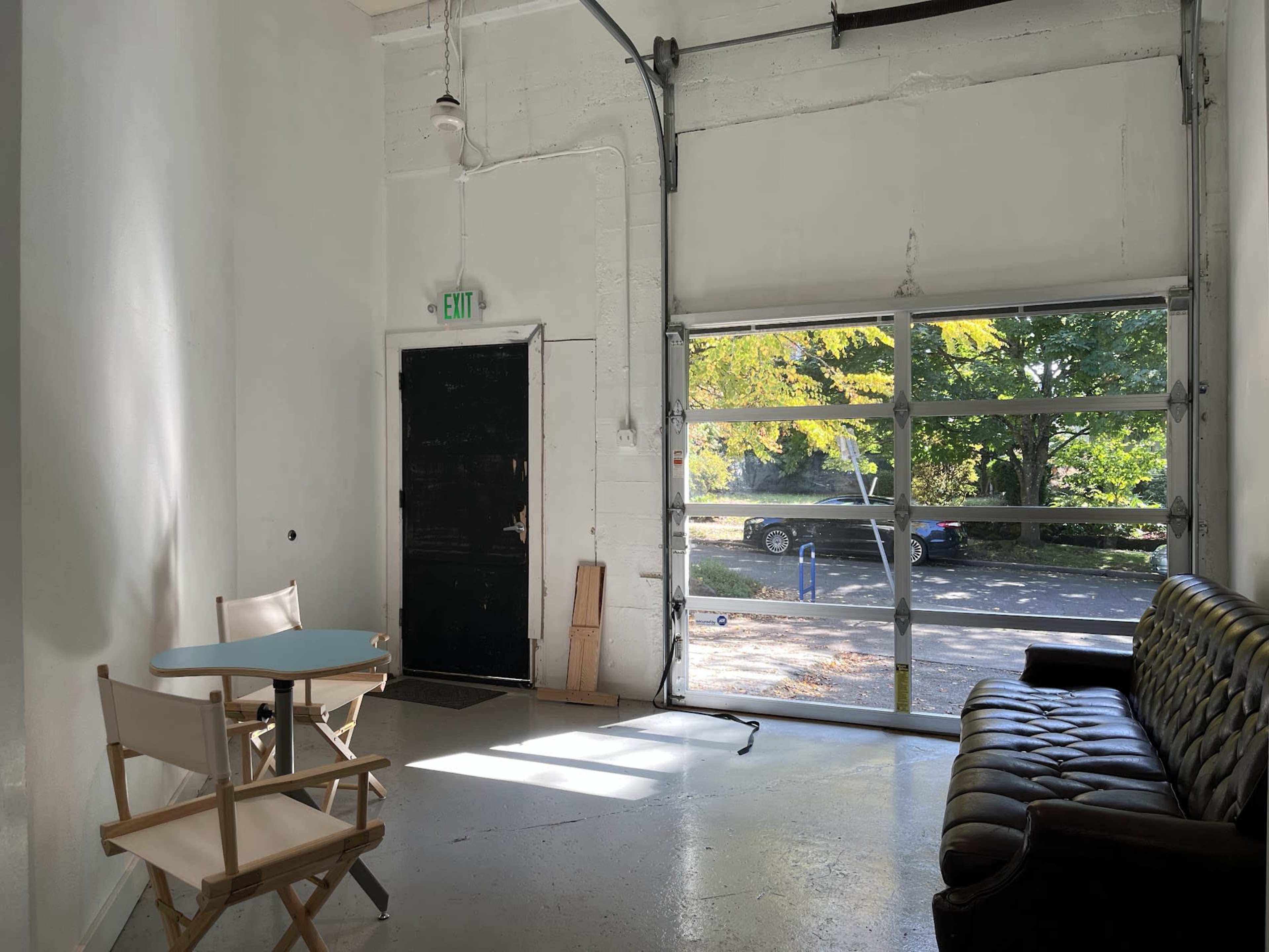 A minimalist interior space with a brown leather sofa and a small round table near a large garage door that opens to an outdoor view.