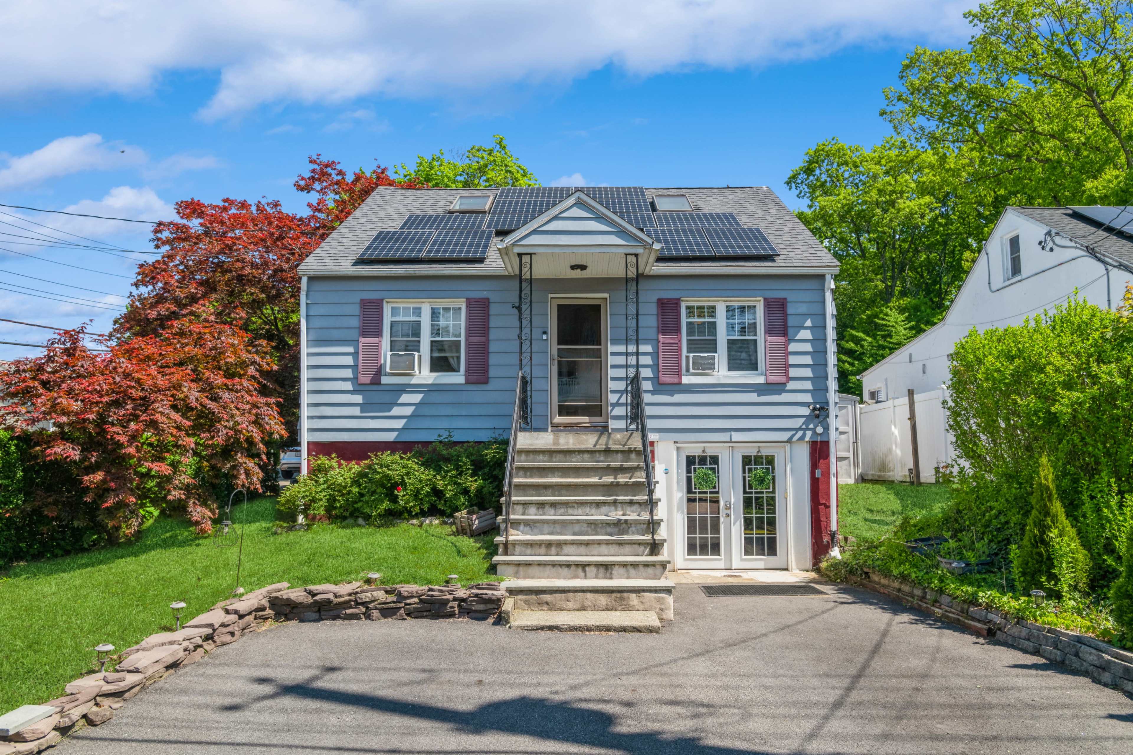 A two-story blue house with a sloped roof features solar panels, red shutters, and a front staircase, surrounded by green grass and landscaped shrubs.