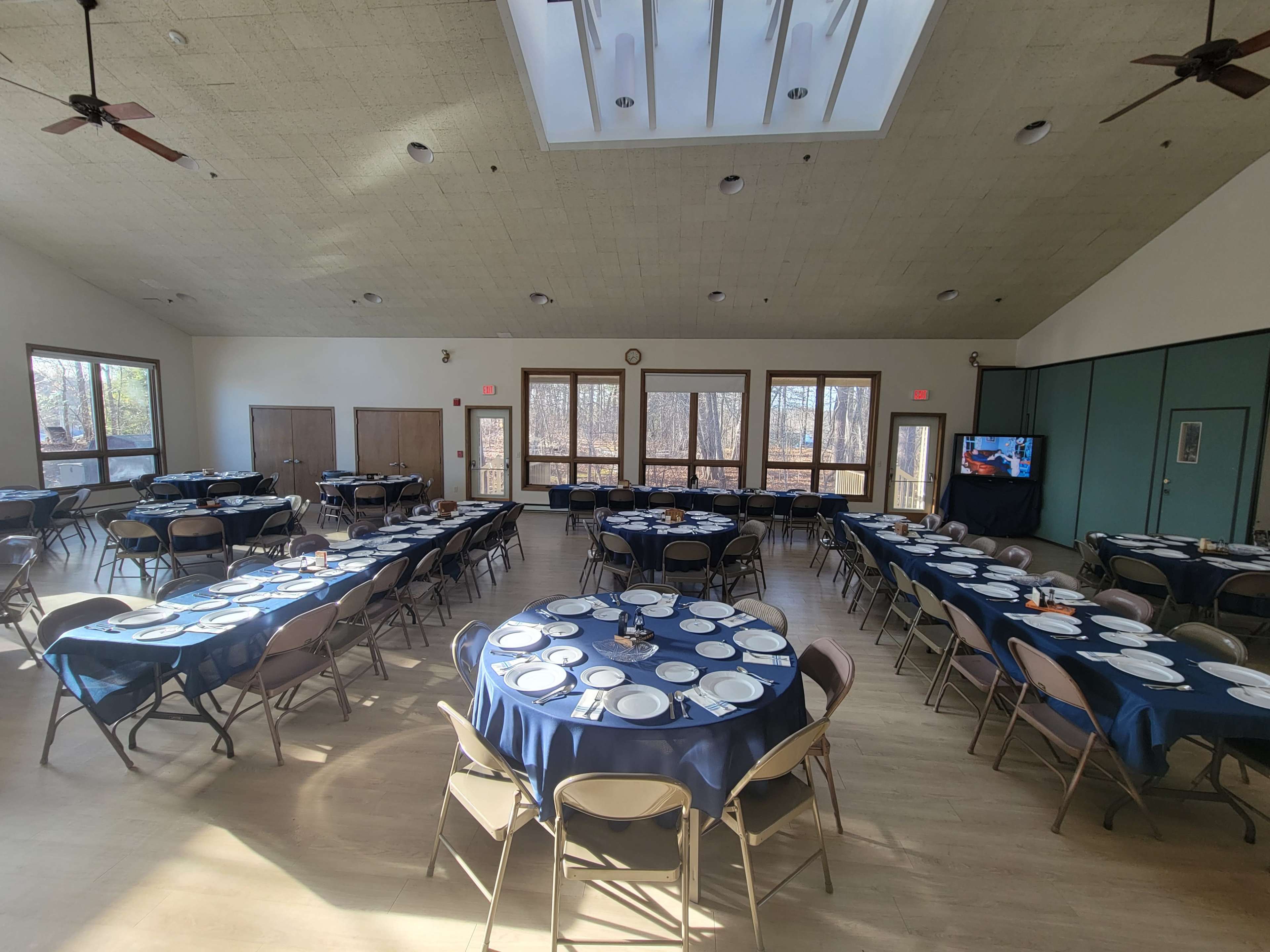 A large meeting room features several round tables set with plates and utensils, arranged for a gathering, with large windows letting in natural light.