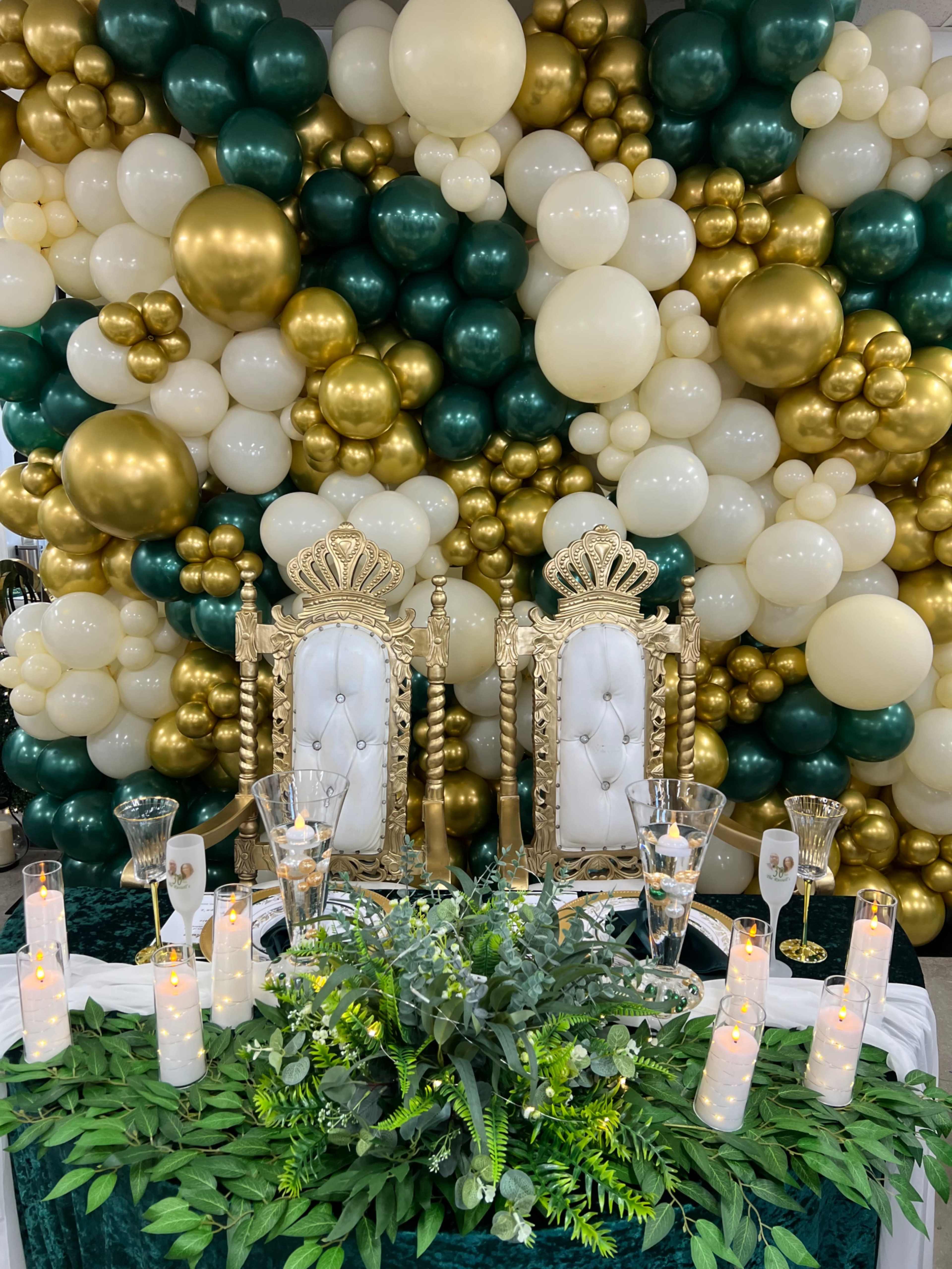 A decorated event space featuring a backdrop of green, white, and gold balloons, with two ornate white thrones and a table adorned with candles and greenery in the foreground.