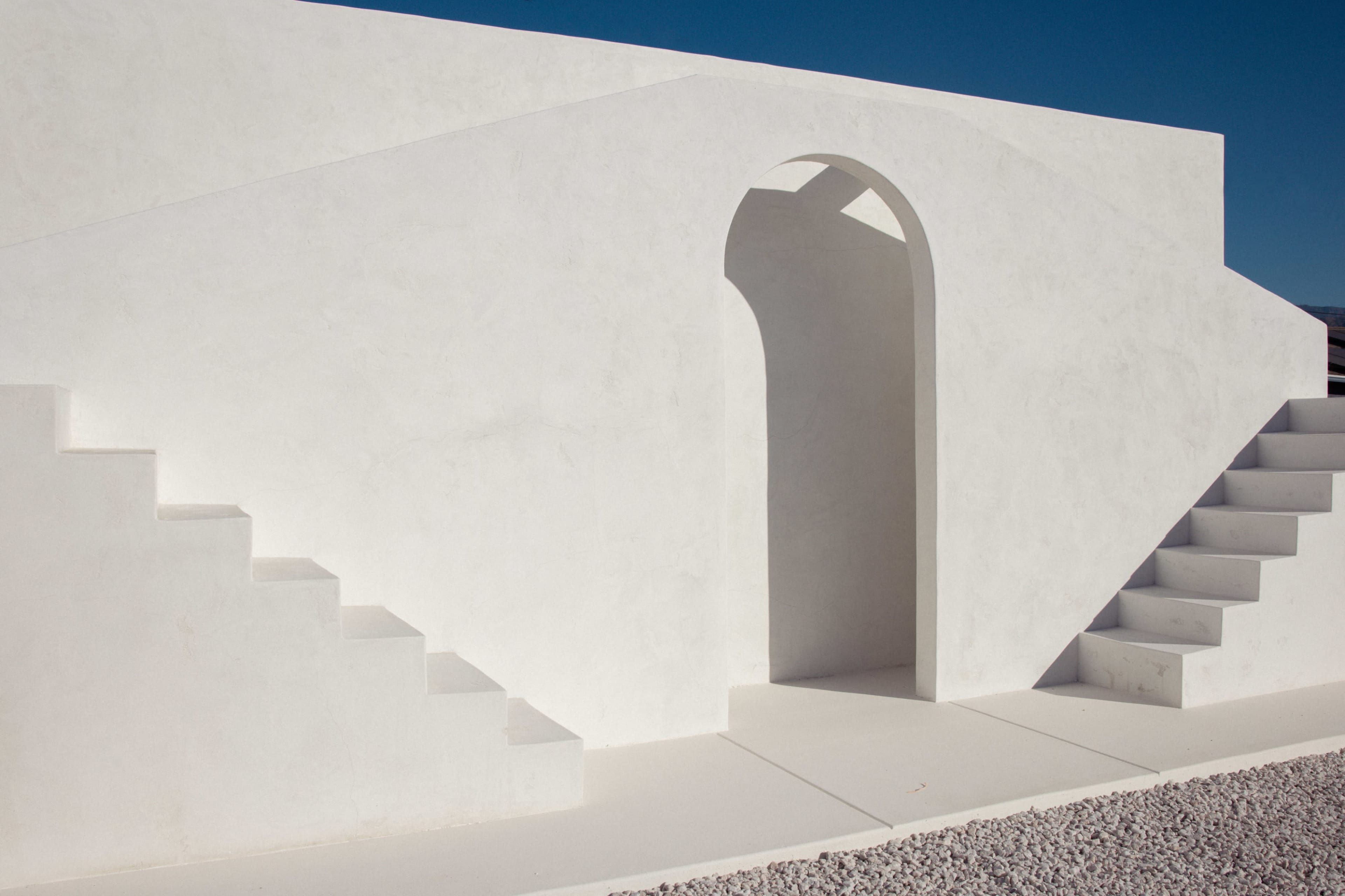 A minimalist architectural structure with white walls, featuring a rounded archway and two sets of stairs.