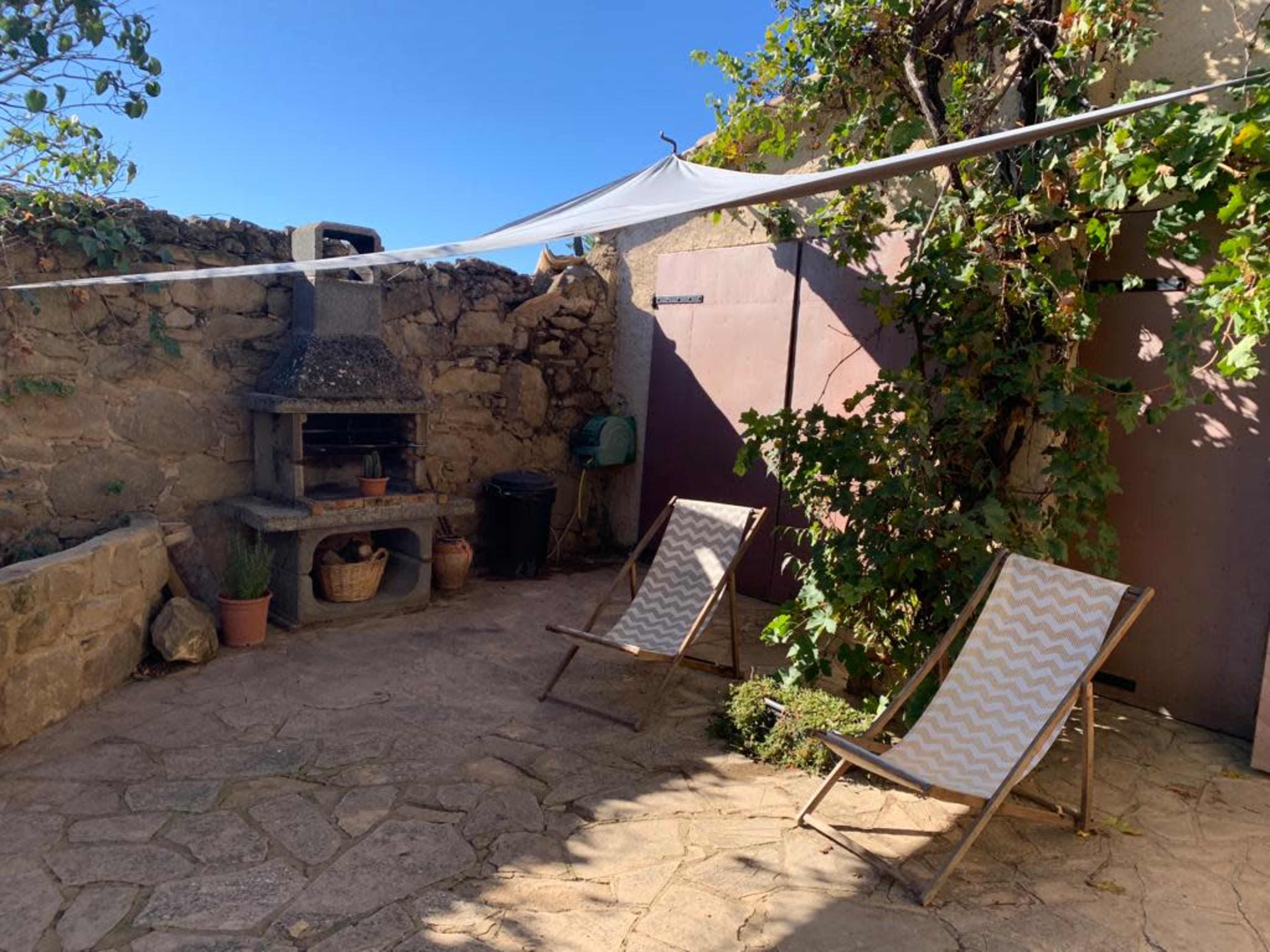 The image shows a stone-paved outdoor area with two deck chairs, a barbecue grill, and climbing plants around a wall.