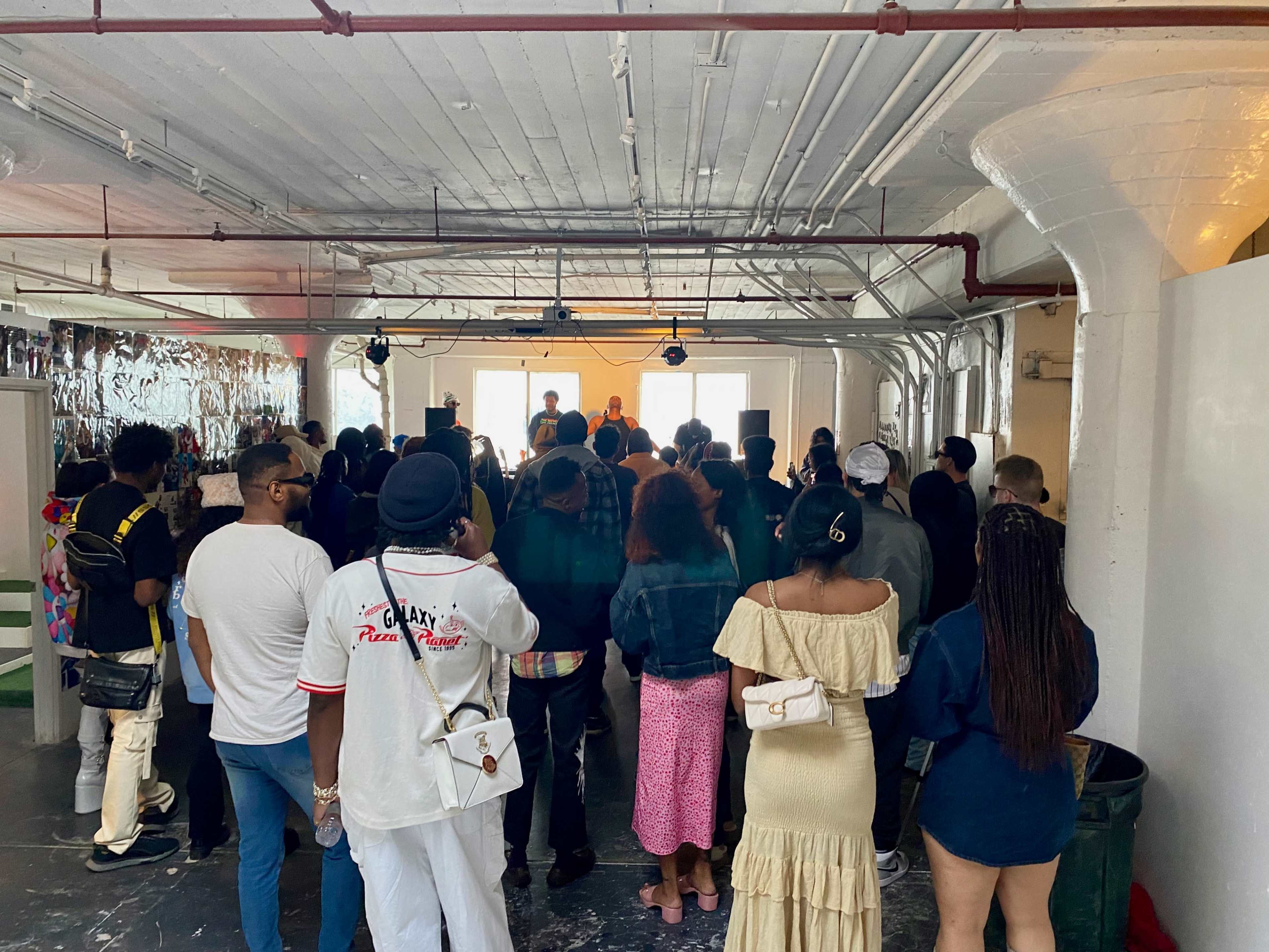 A crowd of people stands in an industrial-style space, listening to a speaker on a small stage.