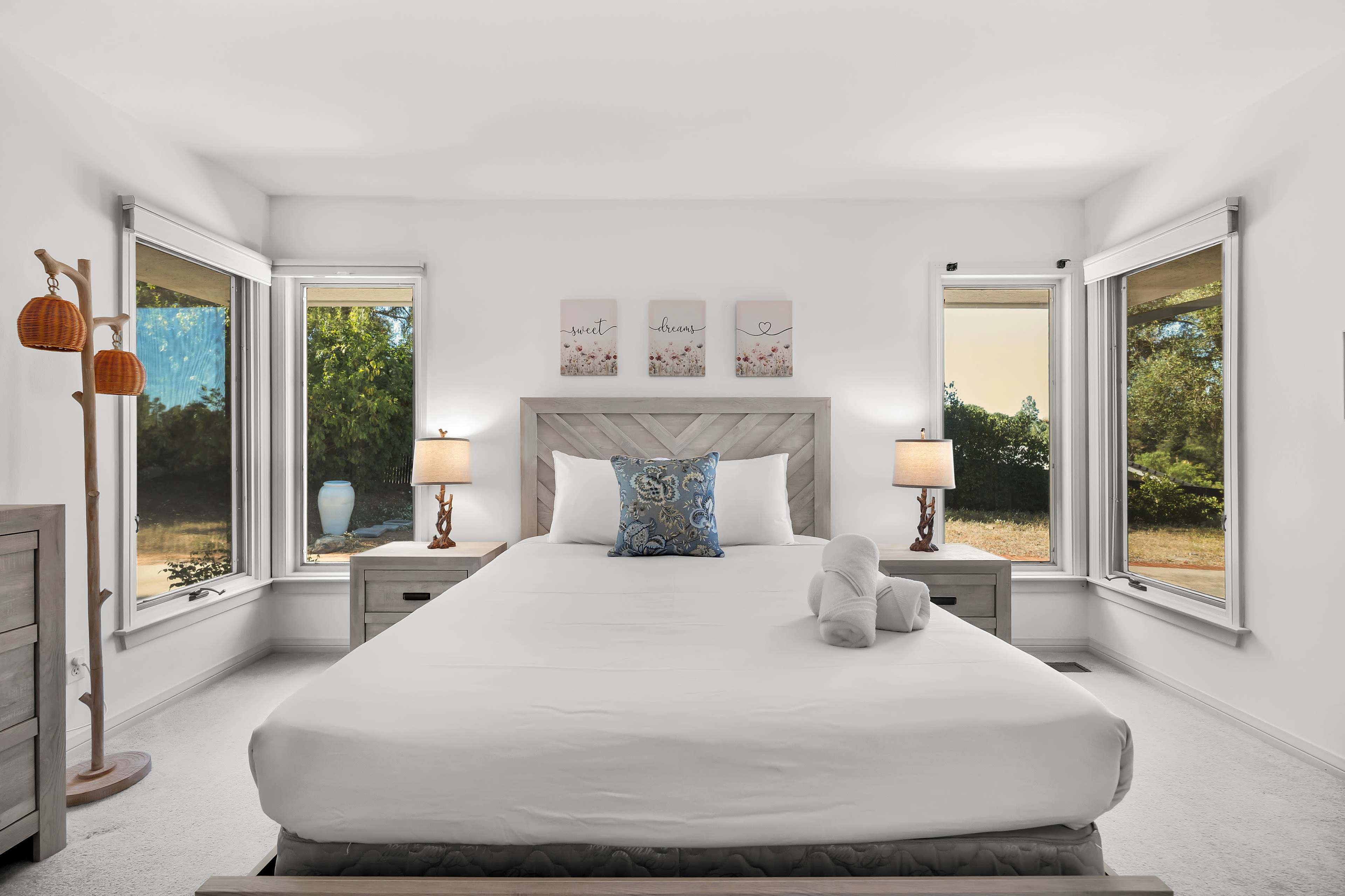 A bright and airy bedroom features a large bed with white bedding, flanked by two nightstands with lamps and windows offering outdoor views.