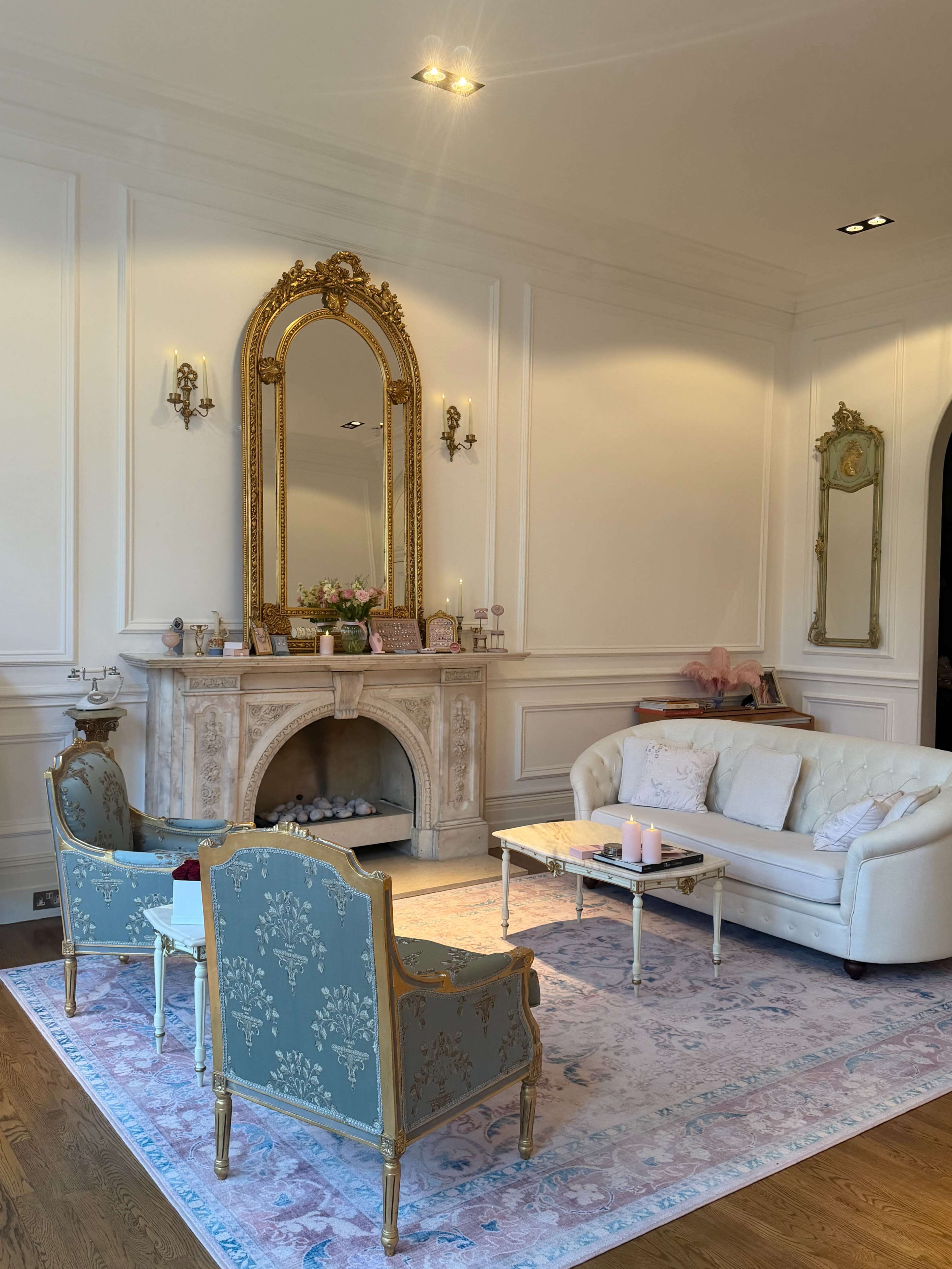 The image shows a elegantly decorated living room featuring a white couch, two ornate teal chairs, a marble fireplace, and a large mirror above it.