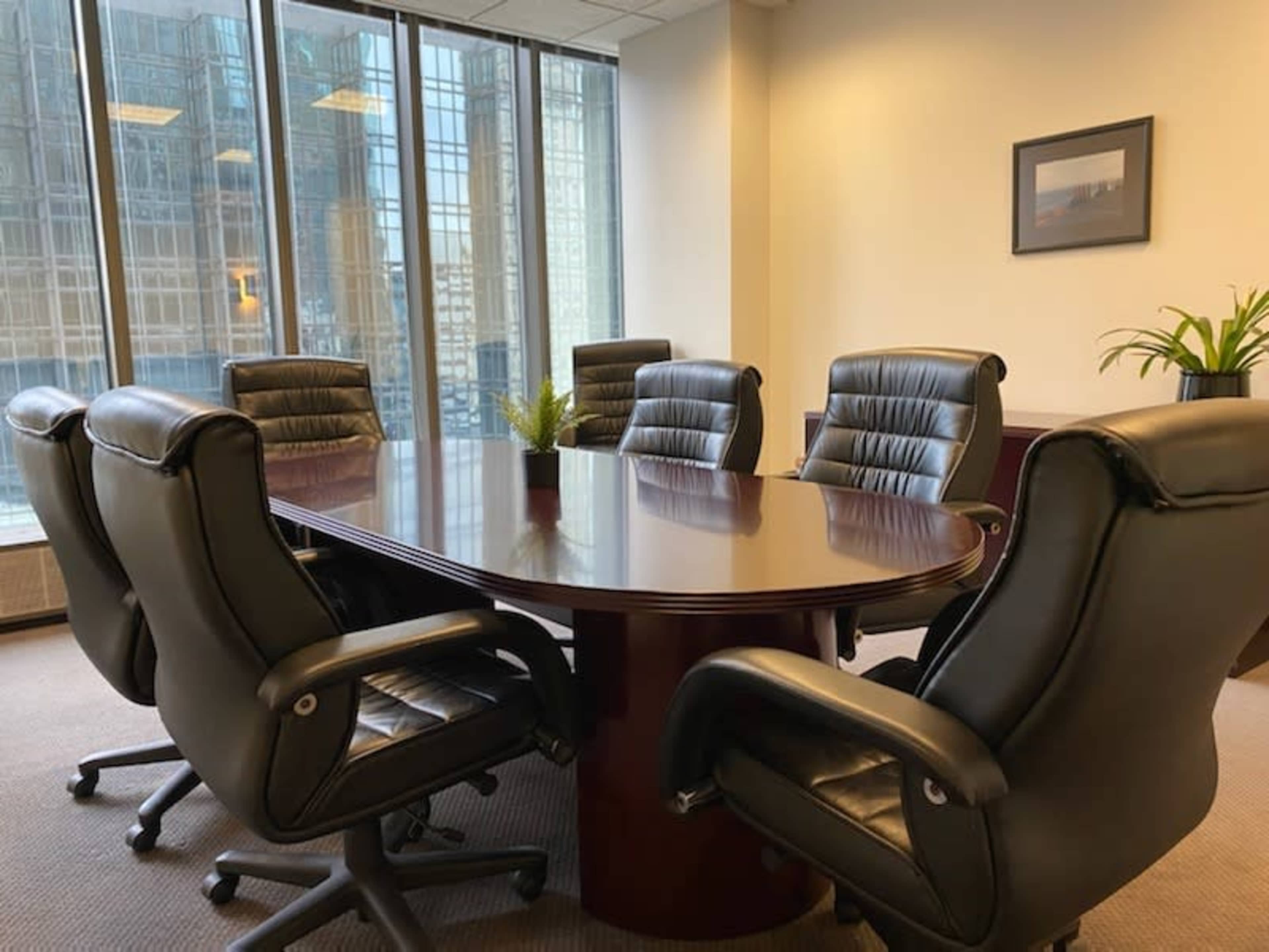 A modern conference room features a large oval table surrounded by eight leather chairs and a small plant on the table.