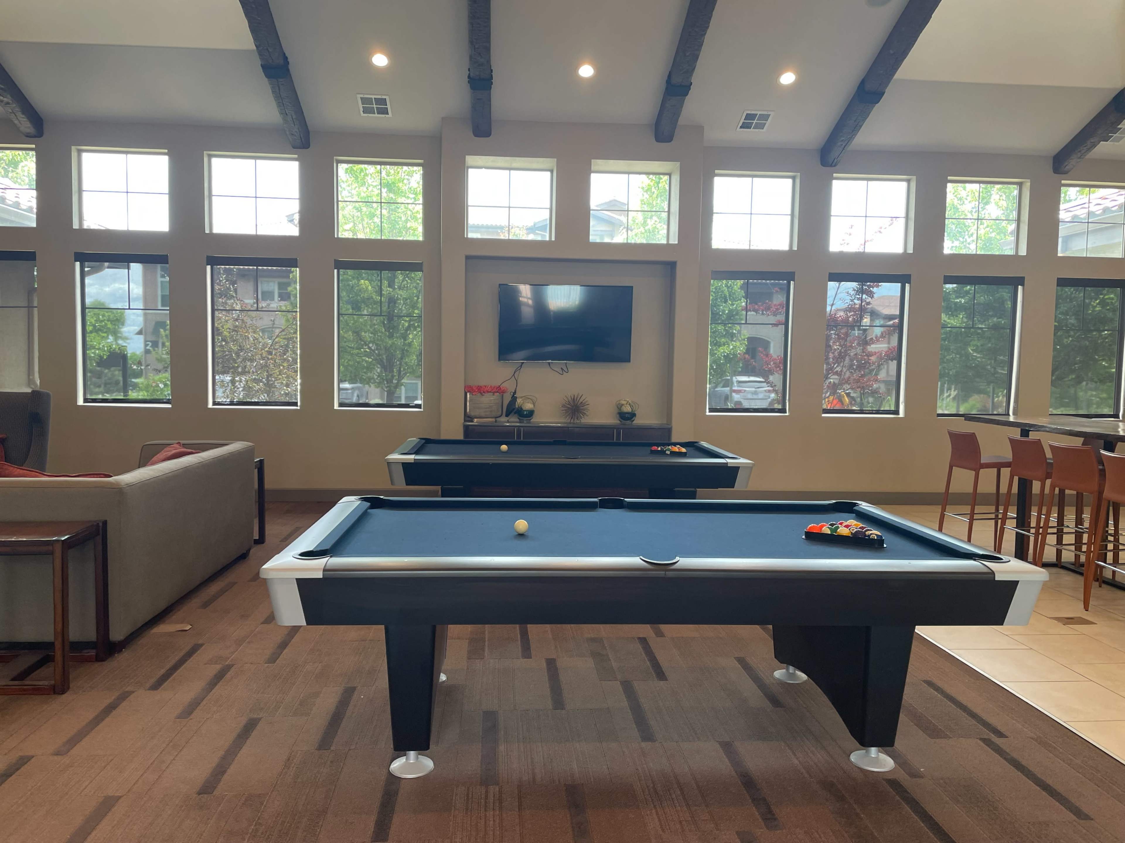 A common room features two pool tables with a television mounted on the wall and large windows allowing natural light to enter.