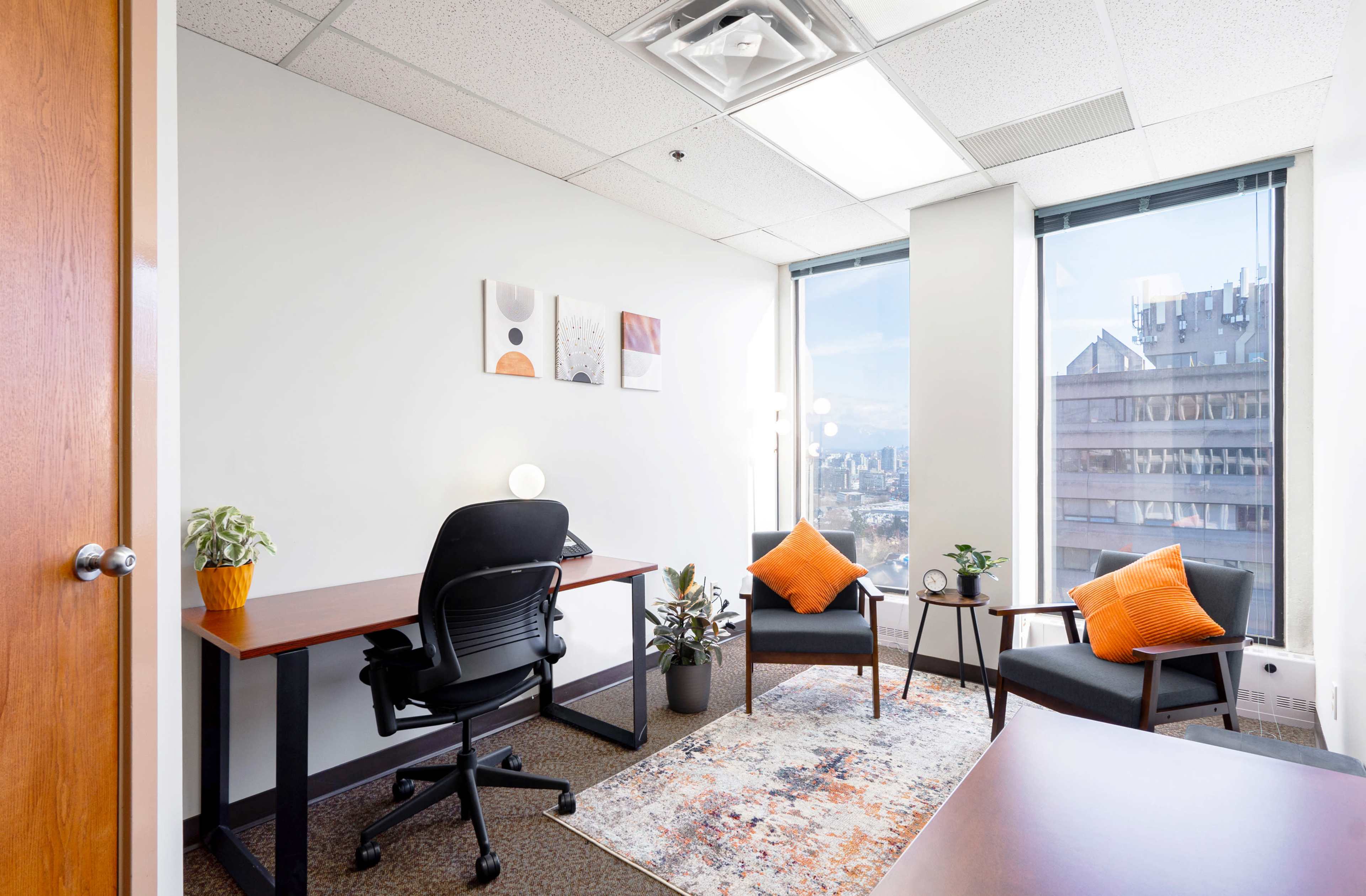 A small office features a desk with a chair, two armchairs with orange cushions, plants, and a rug, all positioned near large windows with a view of a cityscape.