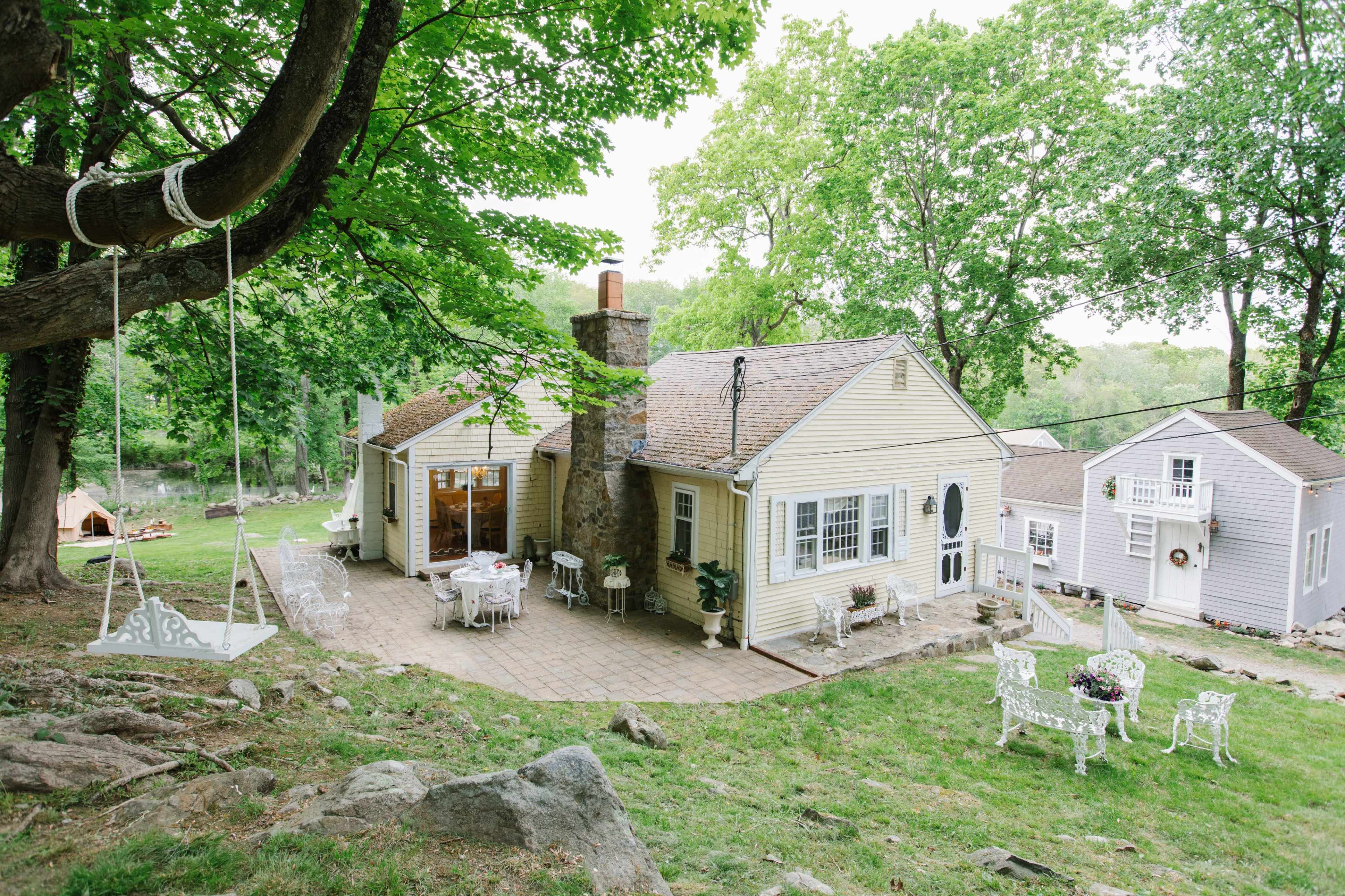 A quaint house with a stone chimney and a patio area surrounded by trees and a swing hangs from a nearby branch.