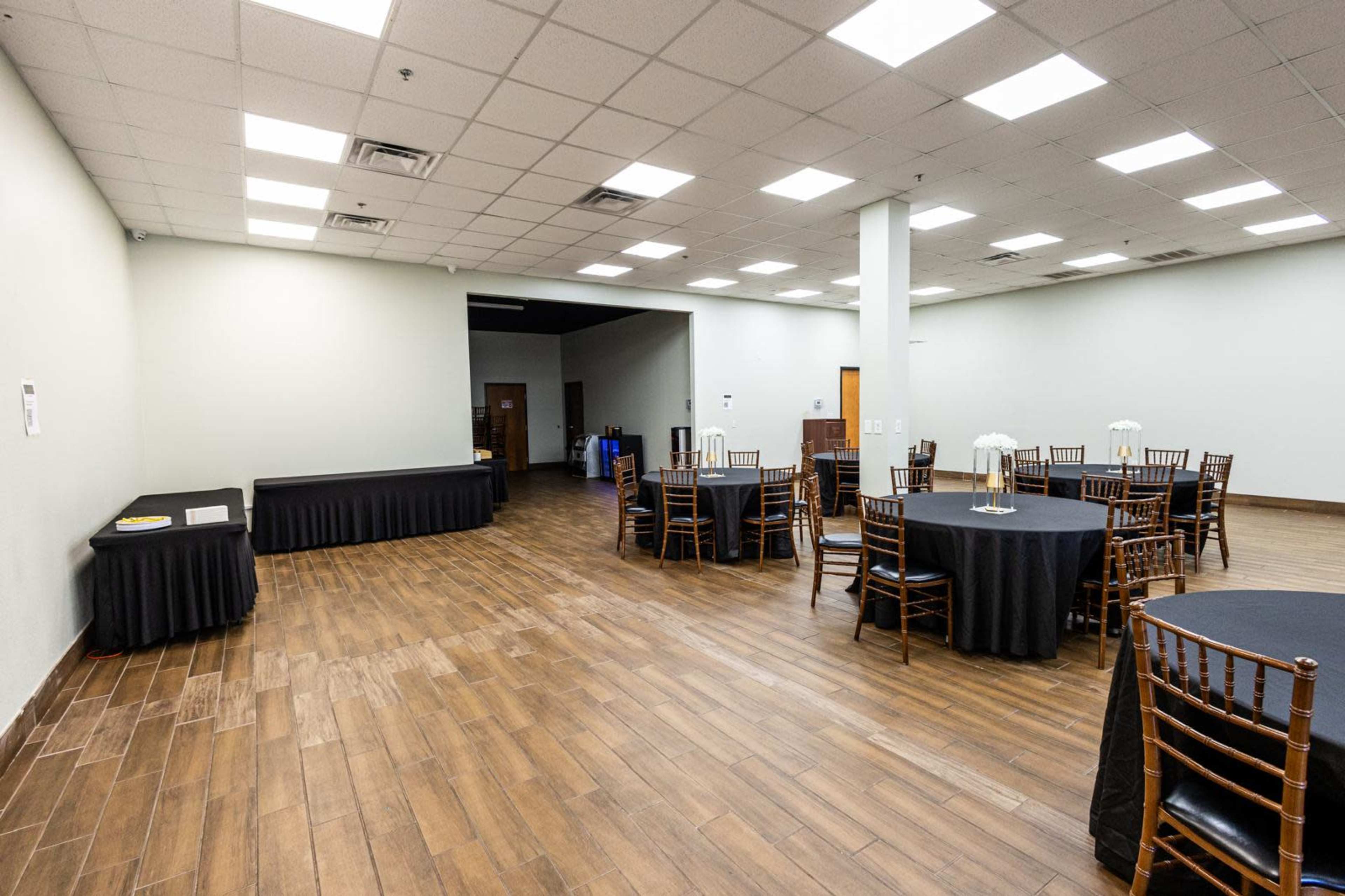 The image shows a spacious event room with several round tables covered in black tablecloths and wooden chairs, along with a black table along one wall.