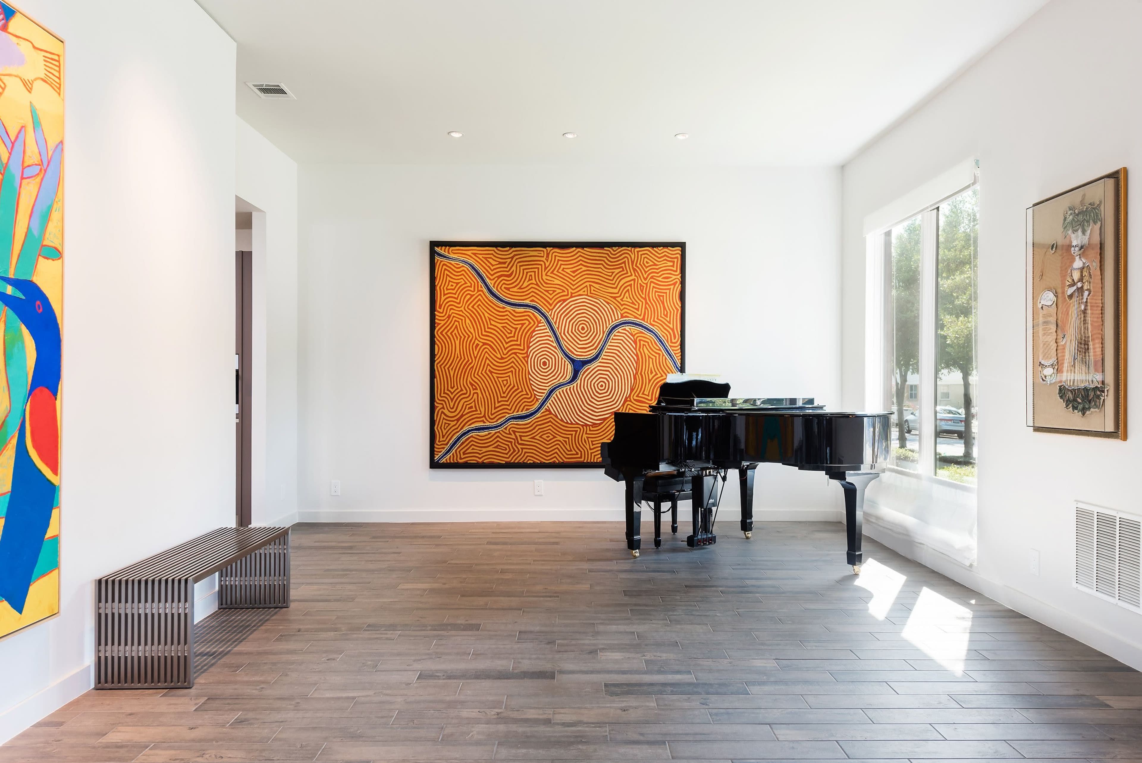 A black grand piano is positioned beside large windows in a room featuring colorful artwork on the walls and hardwood flooring.