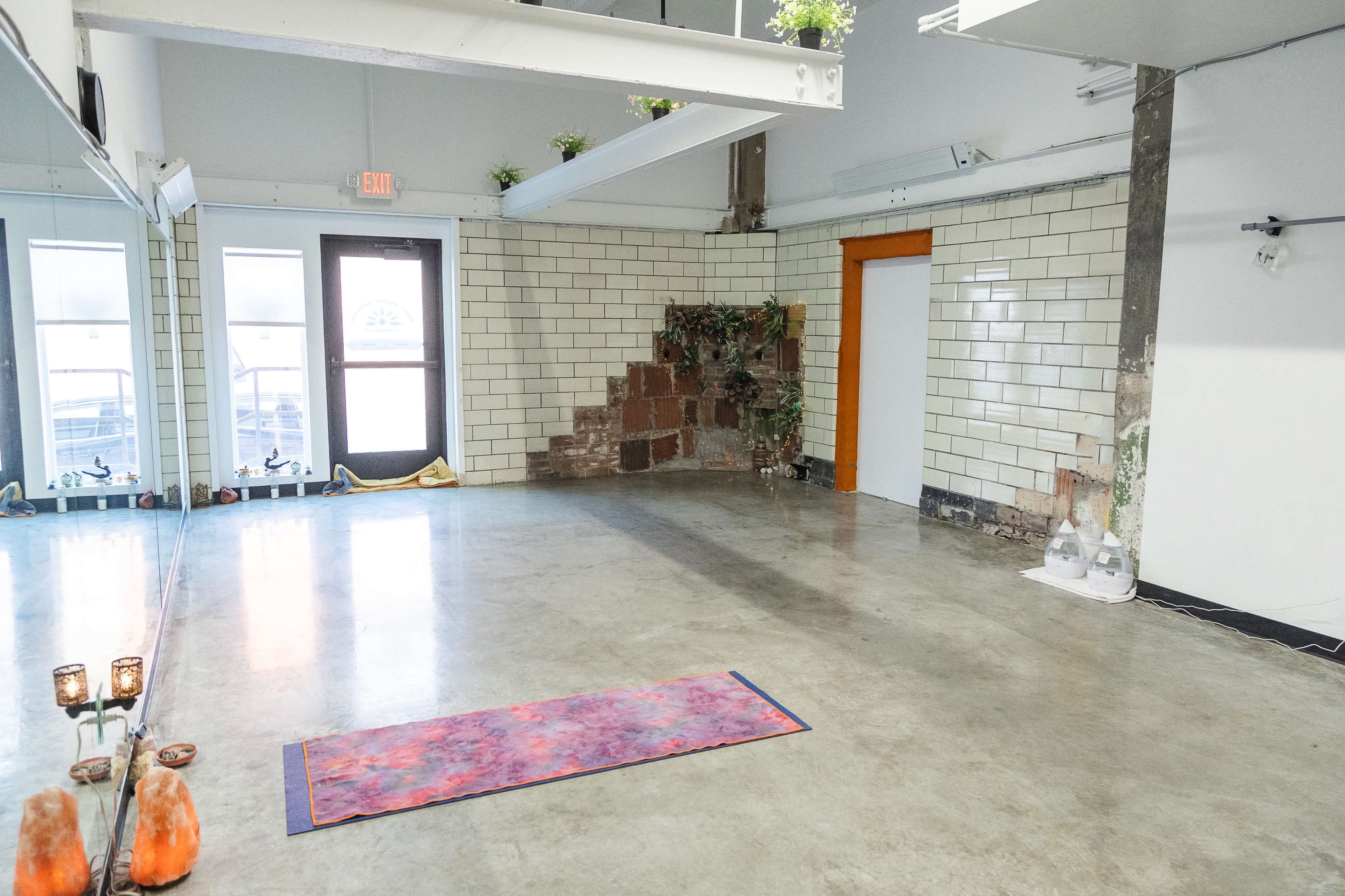 A spacious, minimalist studio with a large mirror, unfinished walls, and a yoga mat placed on the concrete floor.