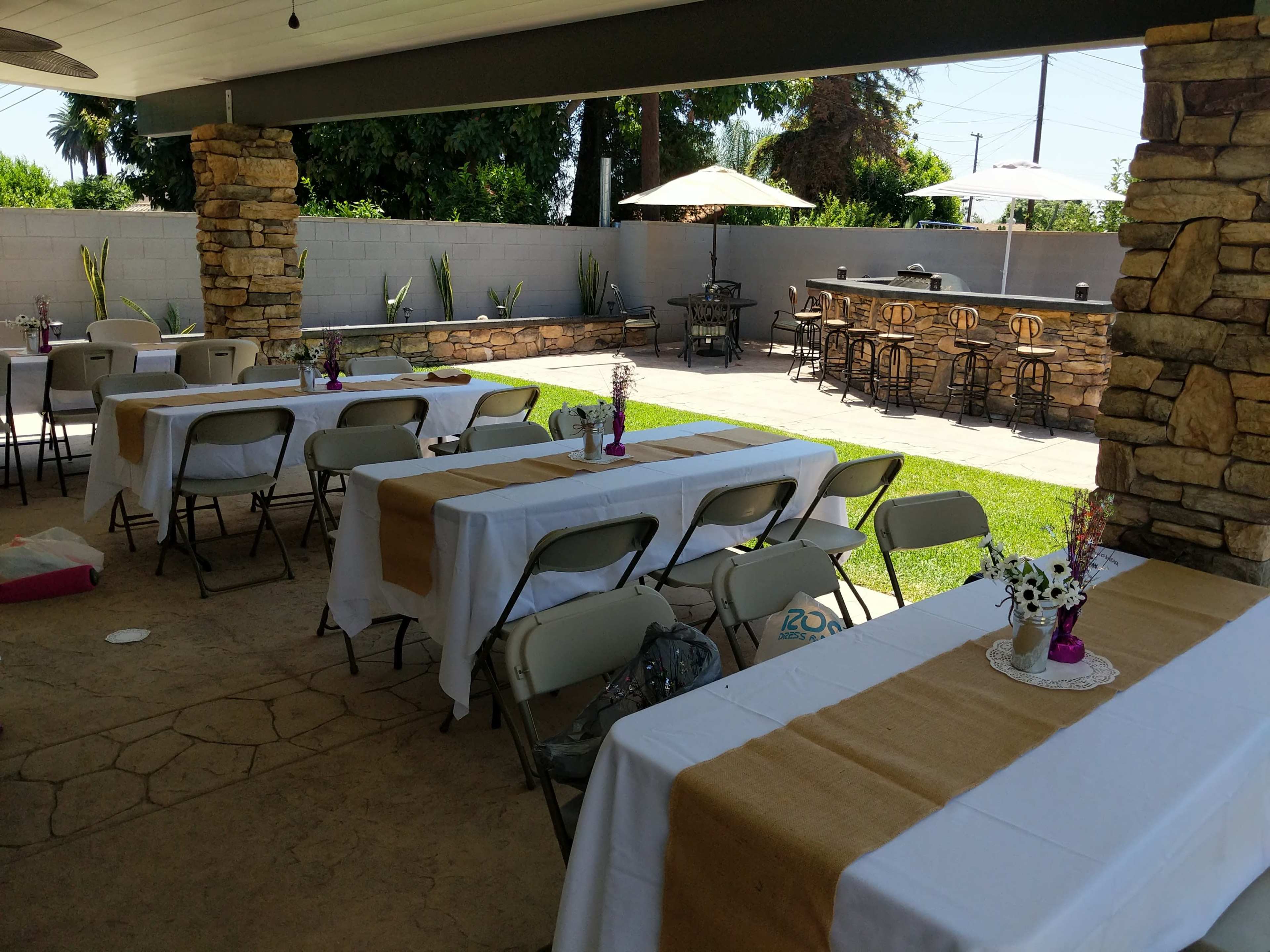 A shaded outdoor space features several tables covered with white tablecloths and brown runners, surrounded by stone columns and greenery, with a bar area in the background.
