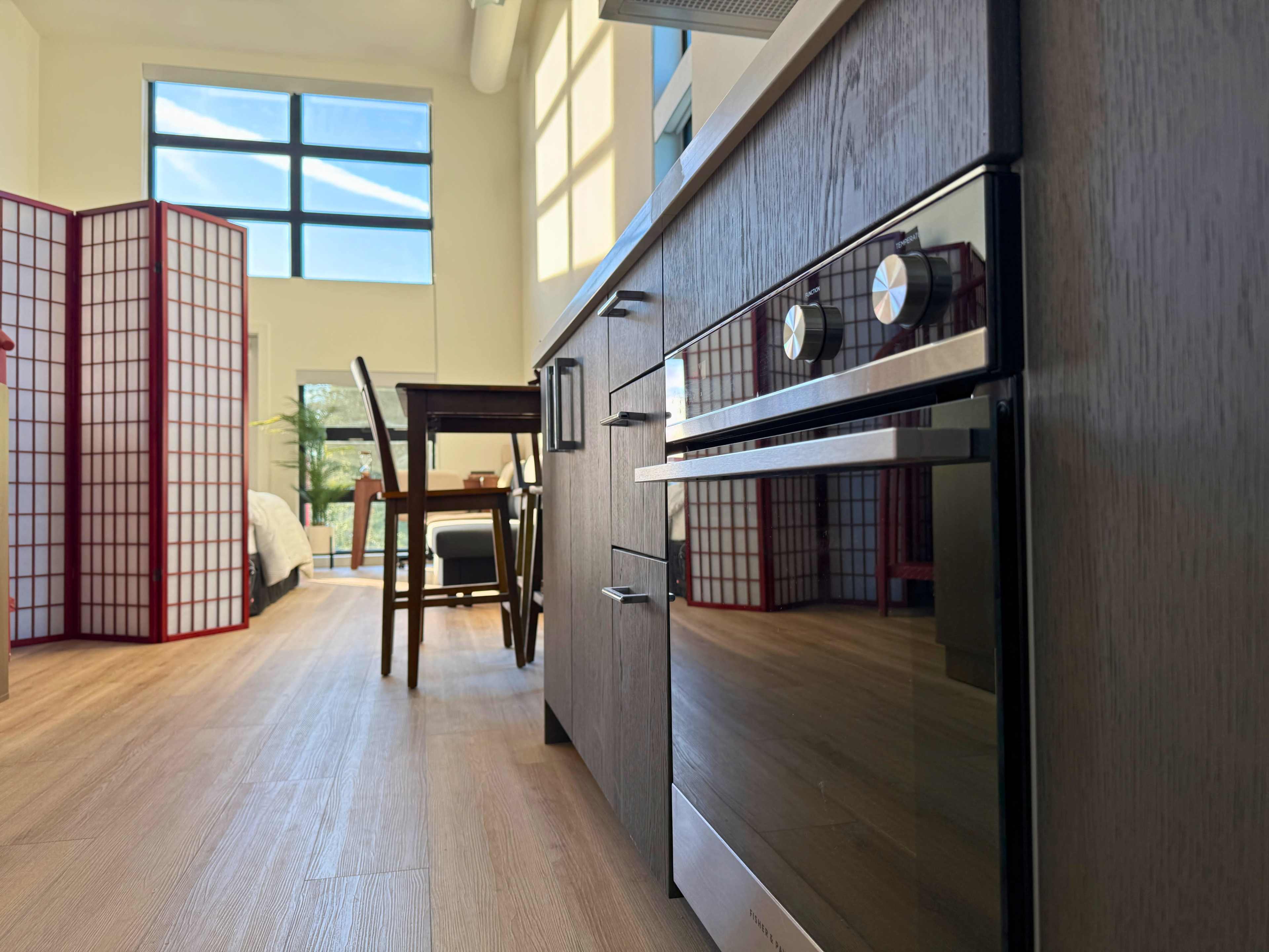 The image shows a modern kitchen space with wooden cabinetry, an oven, and a dining area, all illuminated by large windows.