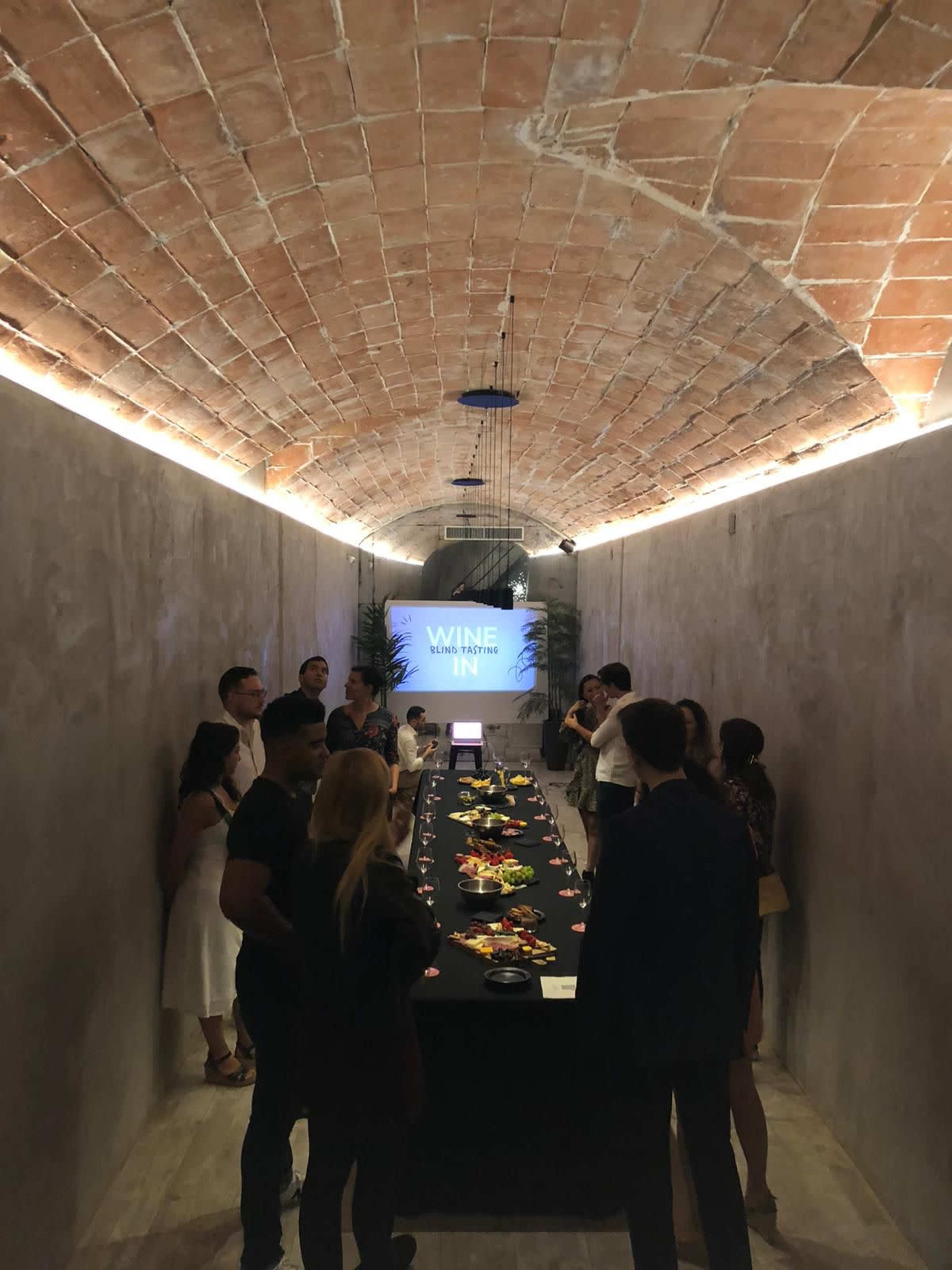 A group of people stands around a long table laden with food in a well-lit, arched room displaying a "WINE IN" sign.