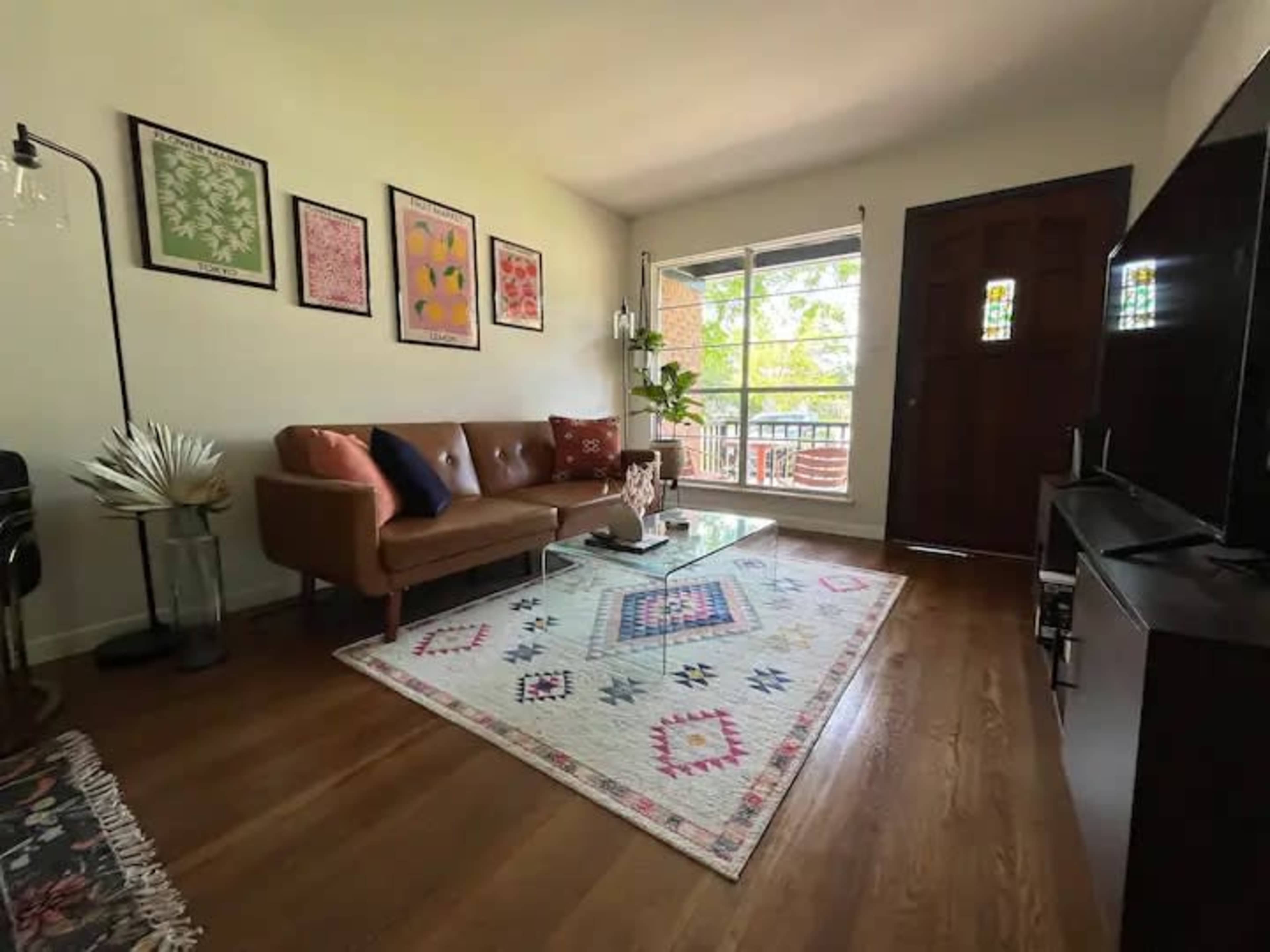 The living room features a brown leather sofa, a glass coffee table, and colorful artwork on the walls, with a large window providing natural light.