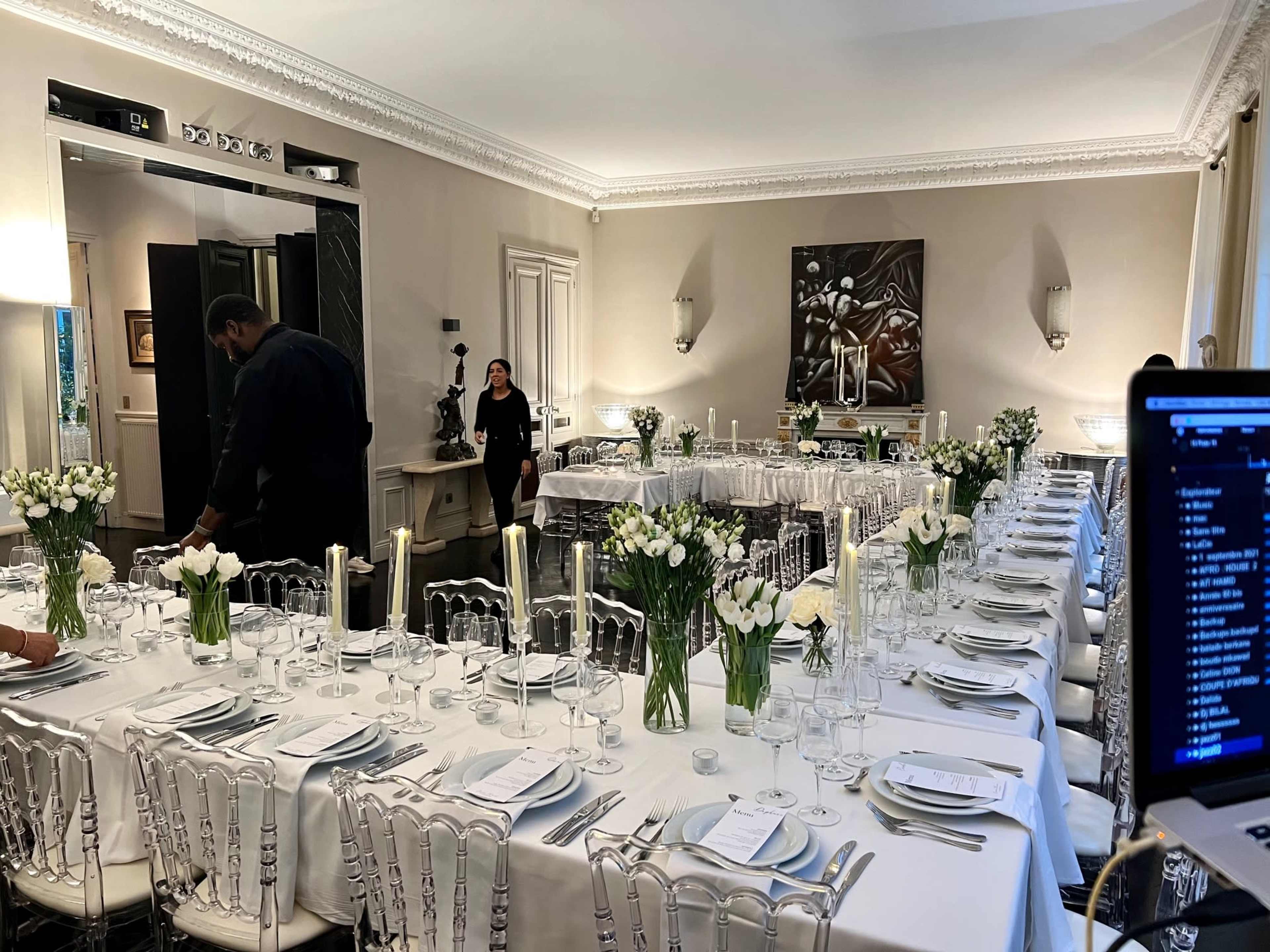 A long, elegantly set dining table with white linens, floral centerpieces, and candle holders is arranged in a well-lit room prepared for an event.