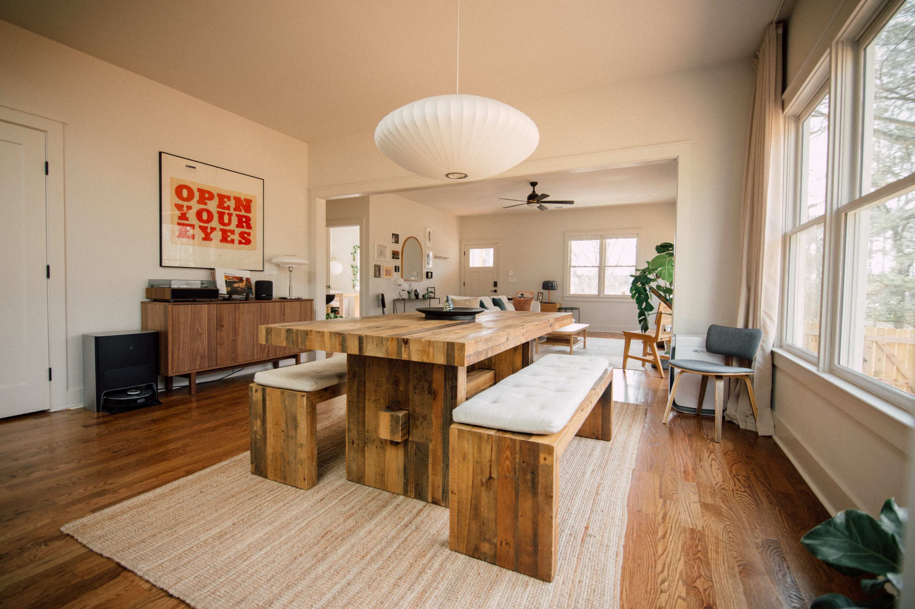 The image features a minimalist dining area with a wooden table and benches, a large pendant light, and large windows allowing natural light to fill the room.