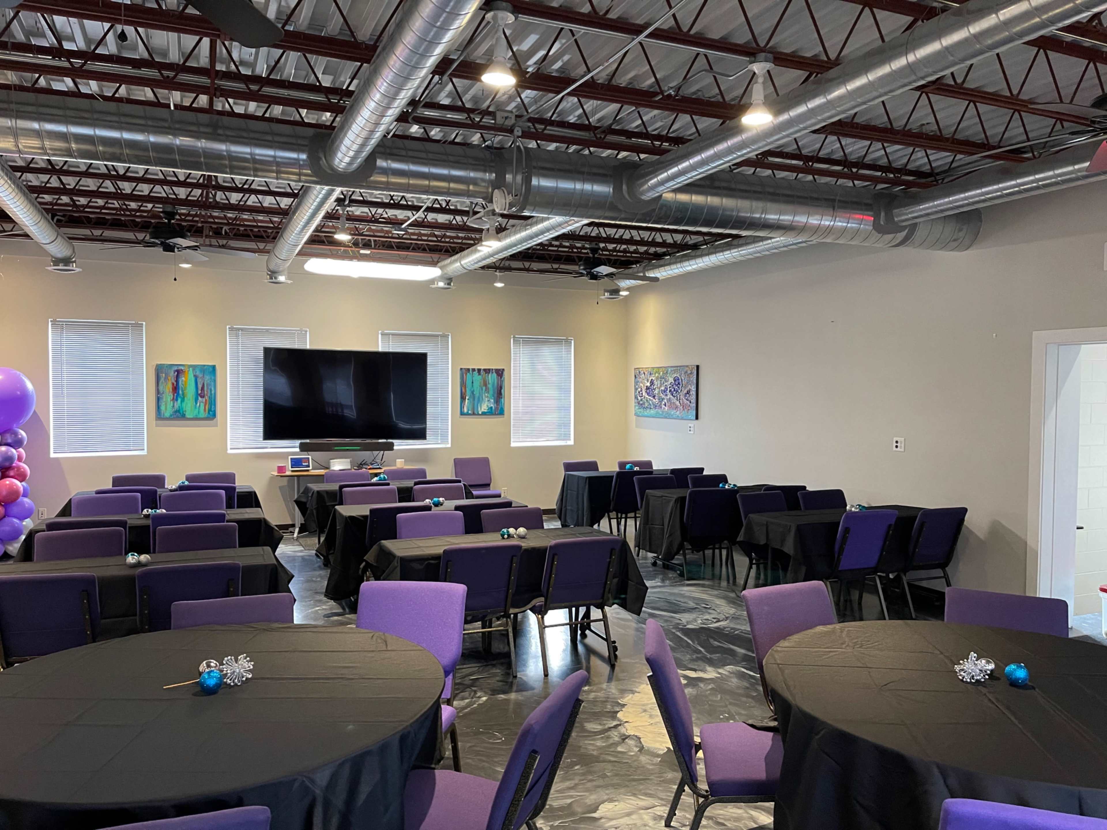 A spacious event room features multiple round black tables with purple chairs, a large screen at the front, and colorful artwork on the walls.
