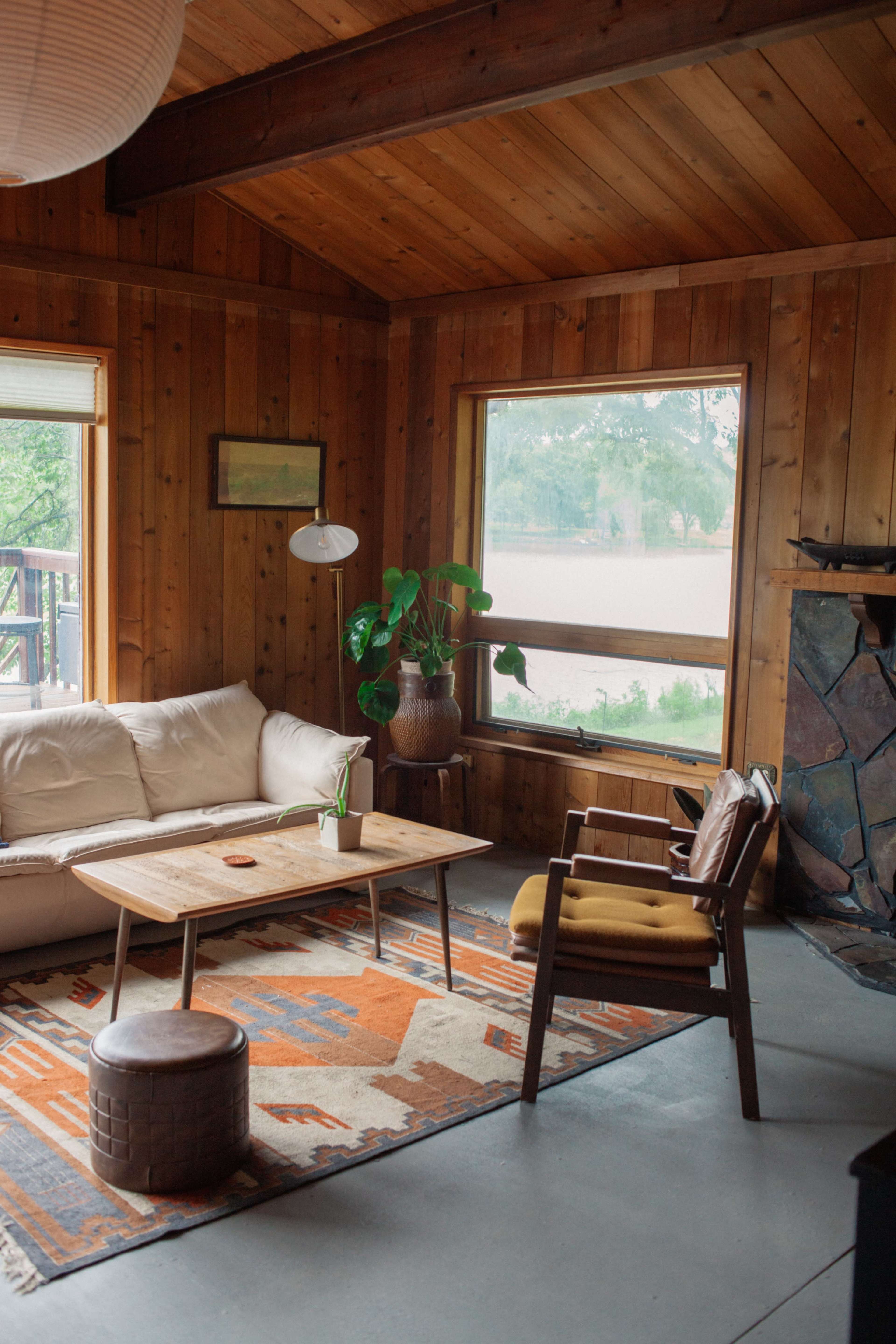 A cozy living room features wood-paneled walls, a light-colored sofa, a wooden coffee table, and a large window with views of greenery outside.