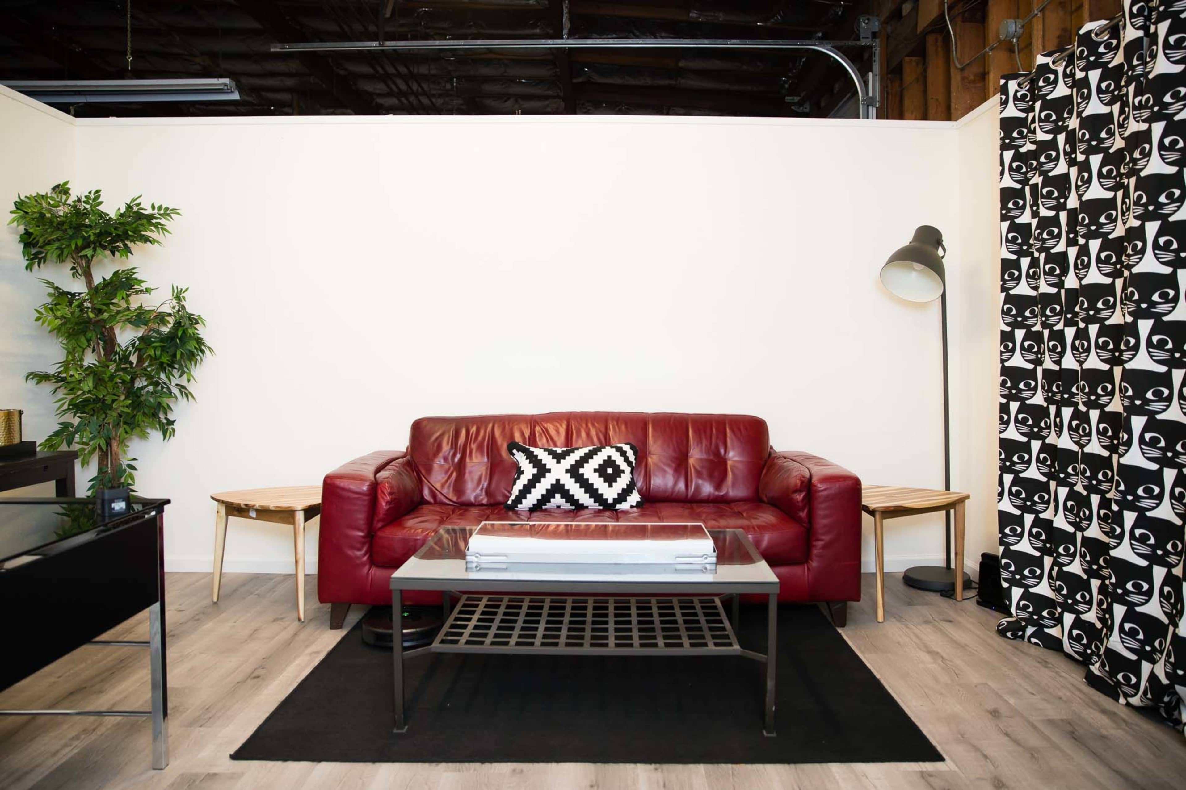 A red leather sofa with a patterned black and white throw pillow, flanked by a small wooden side table and a lamp, in a minimalist room with a wall featuring a black and white graphic design.